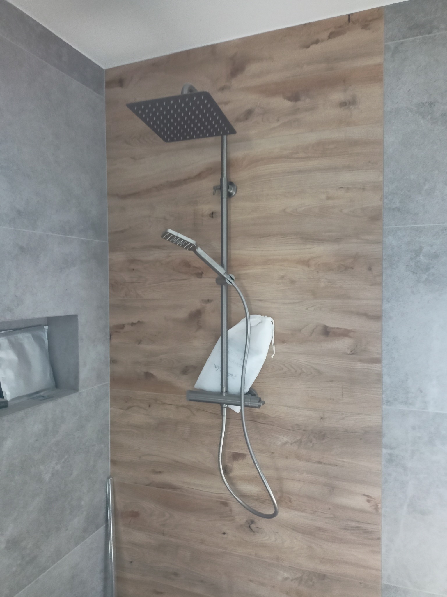 Nowoczesny zestaw prysznicowy z deszczownicą i słuchawką, zamontowany na ścianie wykończonej panelami imitującymi drewno, obok ściany z szarych płytek. W półce ściennej widoczny biały woreczek.