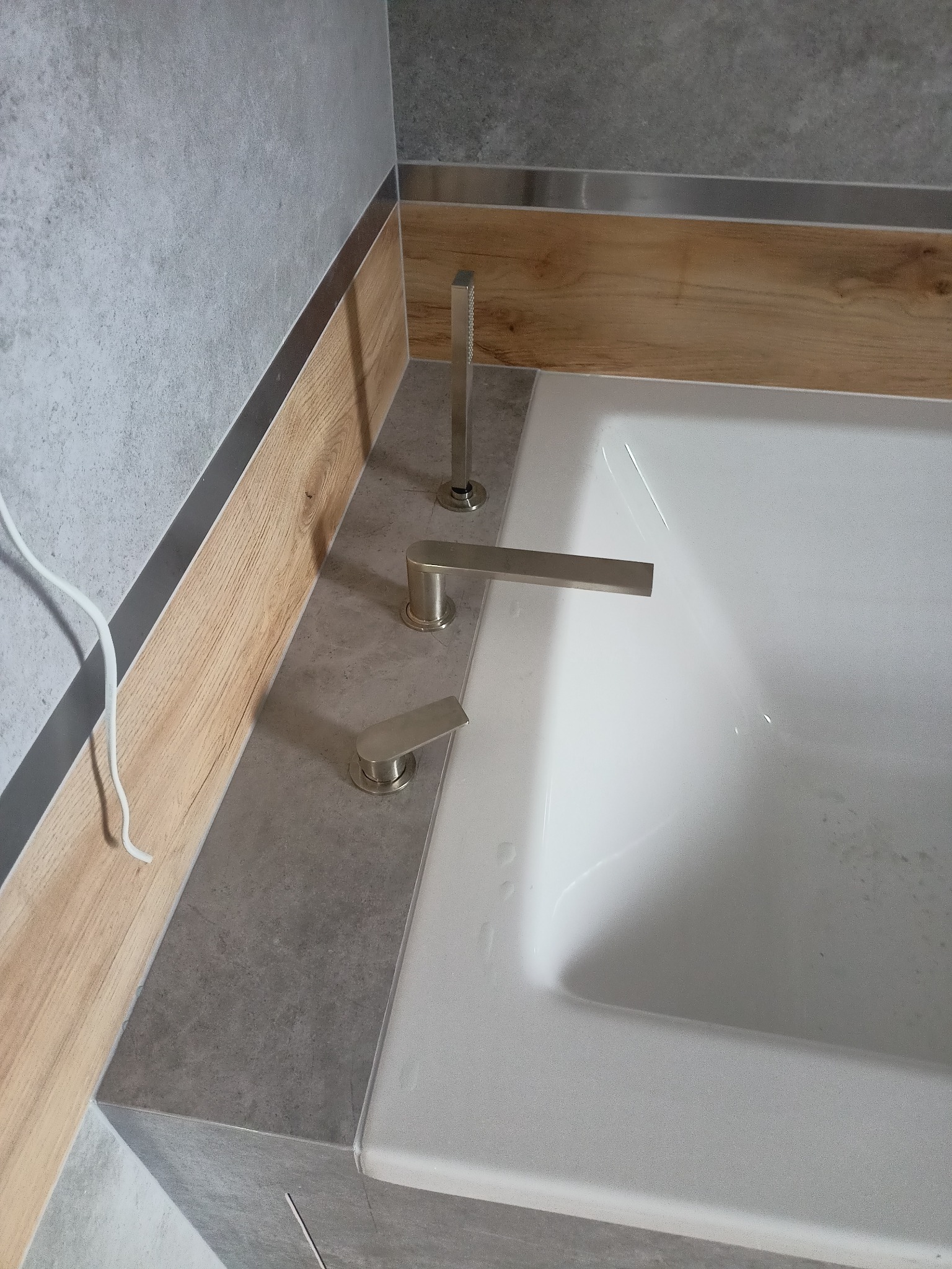 Nowoczesna bateria wannowa w kolorze szczotkowanego niklu zamontowana na obudowie wanny wykończonej szarą płytką, z elementami dekoracyjnymi imitującymi drewno.