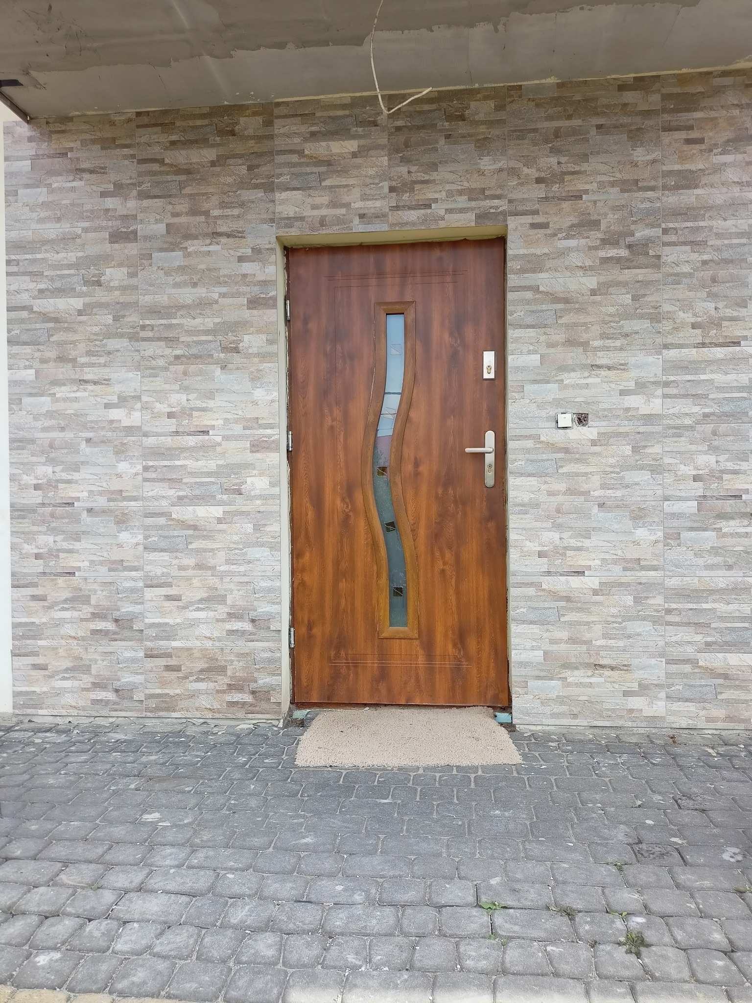 Elewacja budynku z brązowymi drzwiami wejściowymi z szybą o falistym kształcie, wykończona płytkami imitującymi kamień w odcieniach szarości i beżu, widoczny fragment chodnika z kostki brukowej.