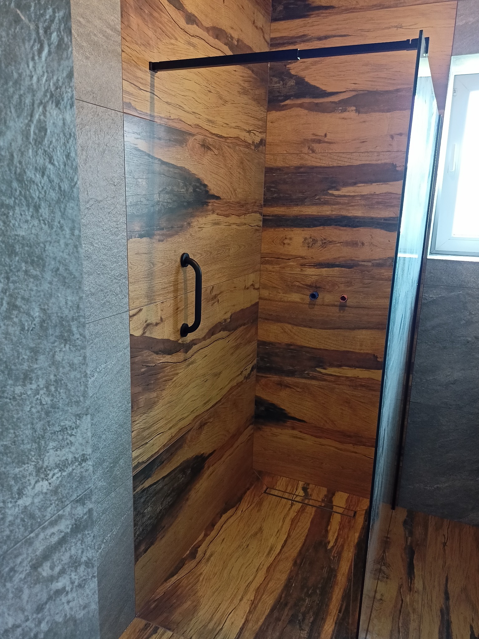 Kabina prysznicowa walk-in z czarnymi profilami i uchwytem, wykończona płytkami imitującymi drewno z widocznymi przyłączami wodnymi.