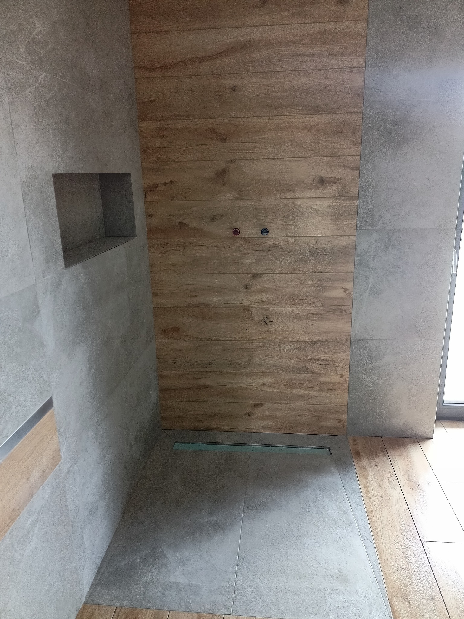 Nowoczesna kabina prysznicowa walk-in z szarymi płytkami imitującymi beton i ścianą z paneli drewnopodobnych, widoczny odpływ liniowy i wnęka w ścianie.