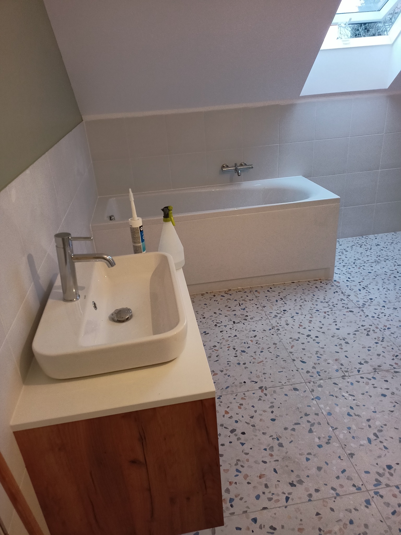 Biała umywalka nablatowa z chromowaną baterią na szafce z drewnopodobnym frontem, w tle wanna obudowana płytkami, szare płytki na ścianach i podłodze z wzorem lastryko, okno dachowe.