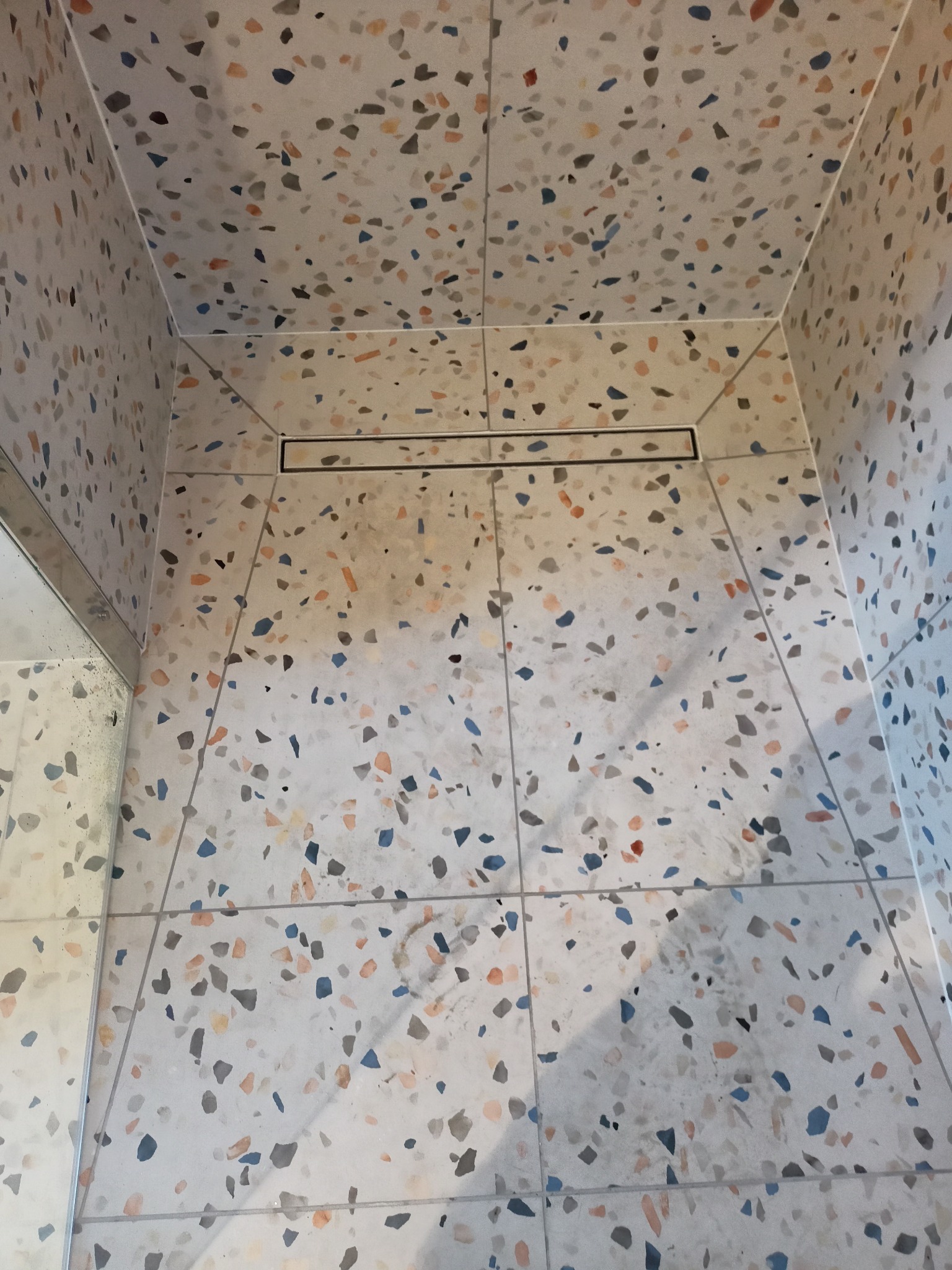 Kabina prysznicowa wykończona płytkami lastryko w odcieniach szarości, błękitu i pomarańczy, z odpływem liniowym w podłodze.