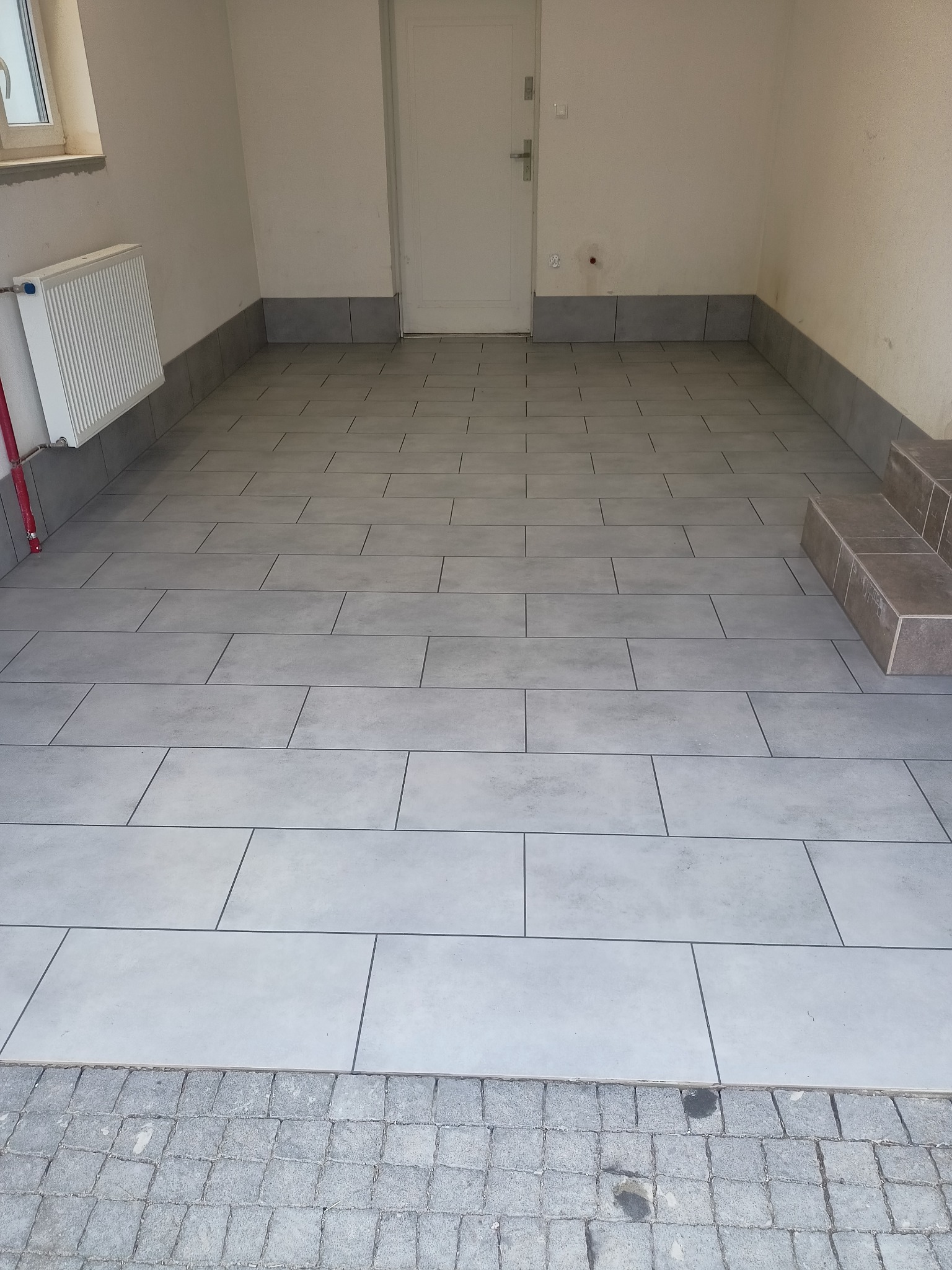 Wyłożona szarymi, prostokątnymi płytkami podłoga w pomieszczeniu, z widocznym grzejnikiem, schodami i fragmentem kostki brukowej przy progu.