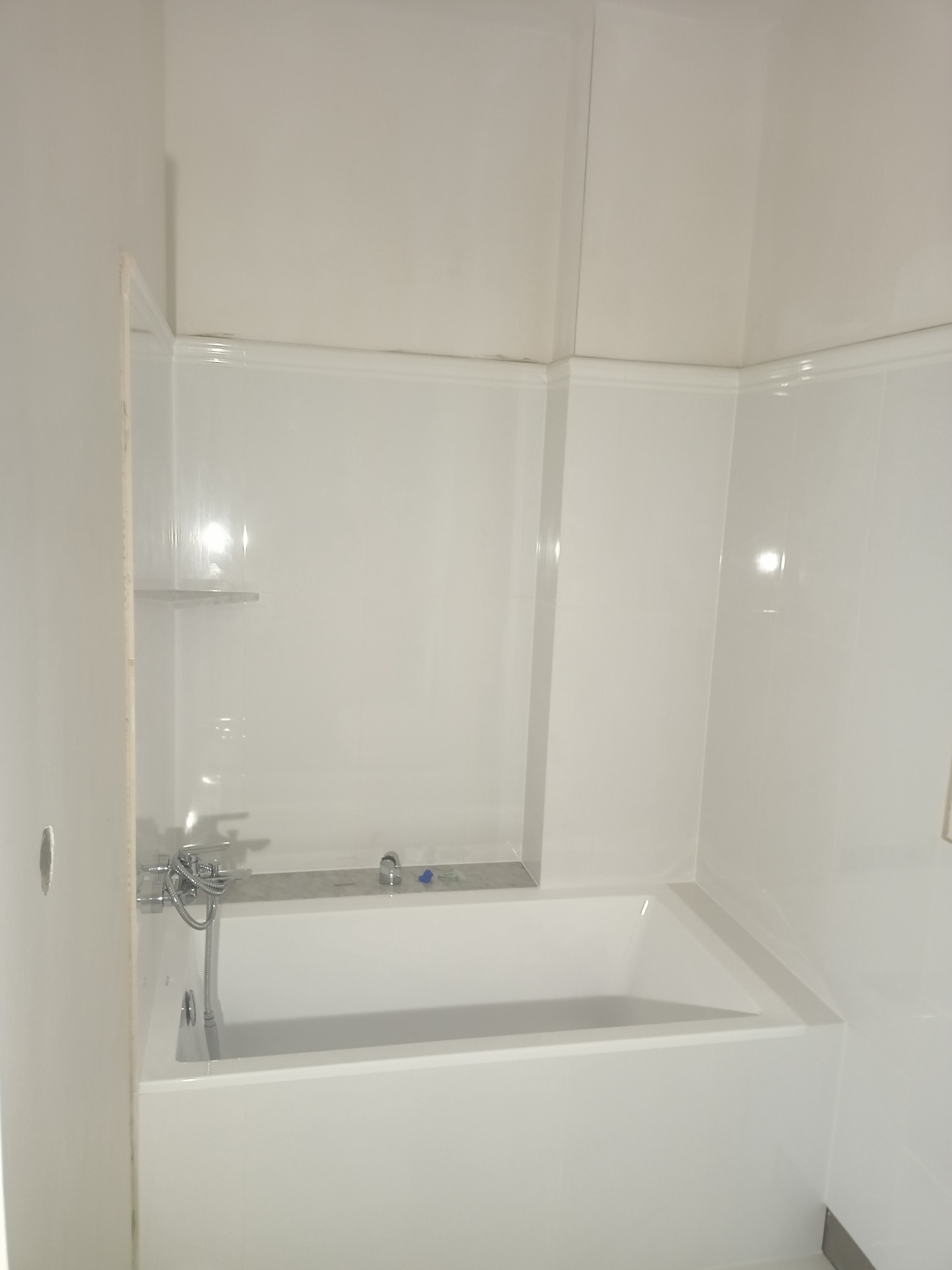 Biała, wykończona wanna w obudowie z białych paneli ściennych, widoczna bateria wannowa z słuchawką prysznicową oraz niewielka półka ścienna.