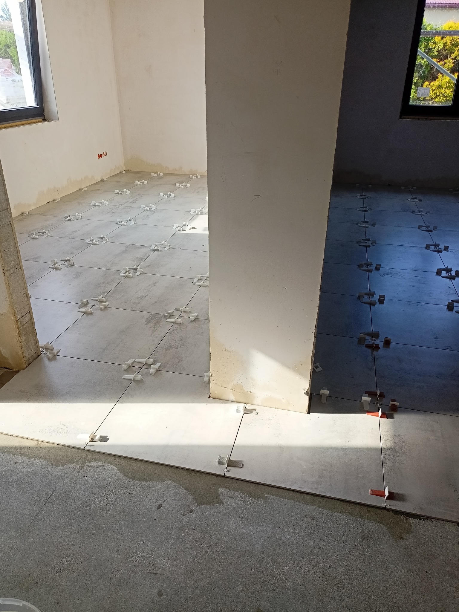 Układanie szarych płytek podłogowych w dwóch pomieszczeniach oddzielonych filarem, z użyciem białych i czerwonych krzyżyków dystansowych, widoczne surowe ściany i podłoże.