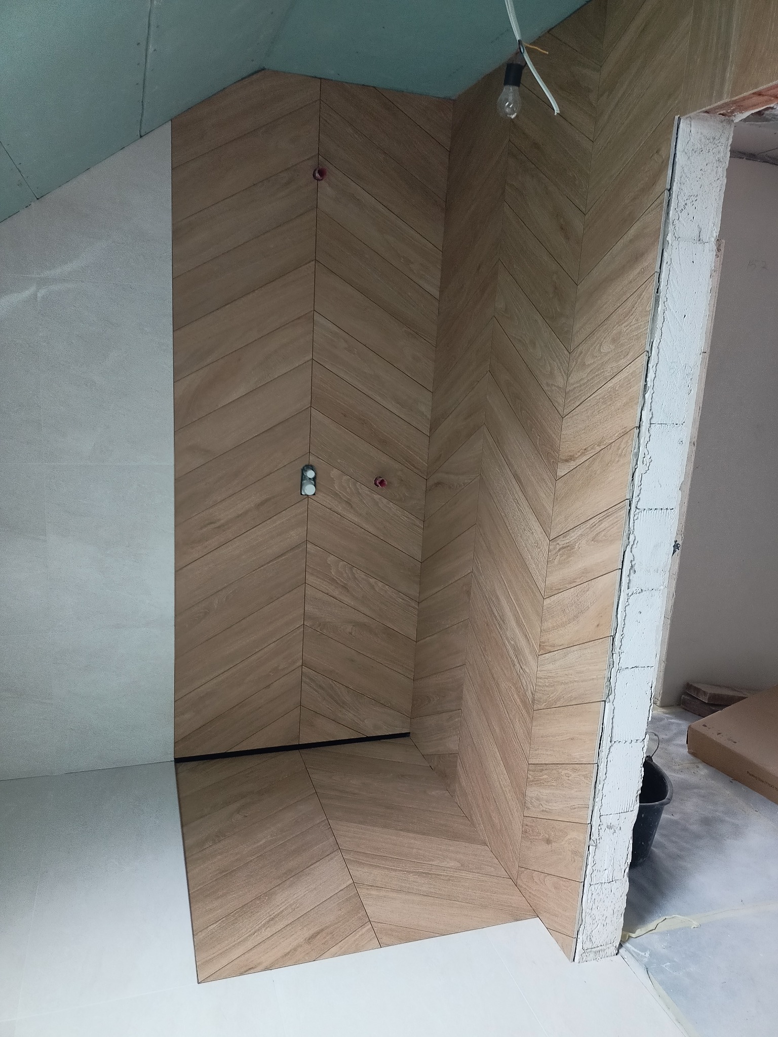 Ukończona ściana prysznicowa z płytek ułożonych w jodełkę w odcieniach drewna, kontrastująca z jasnymi płytkami na sąsiedniej ścianie i podłodze, widoczne wystające instalacje, surowy stan...