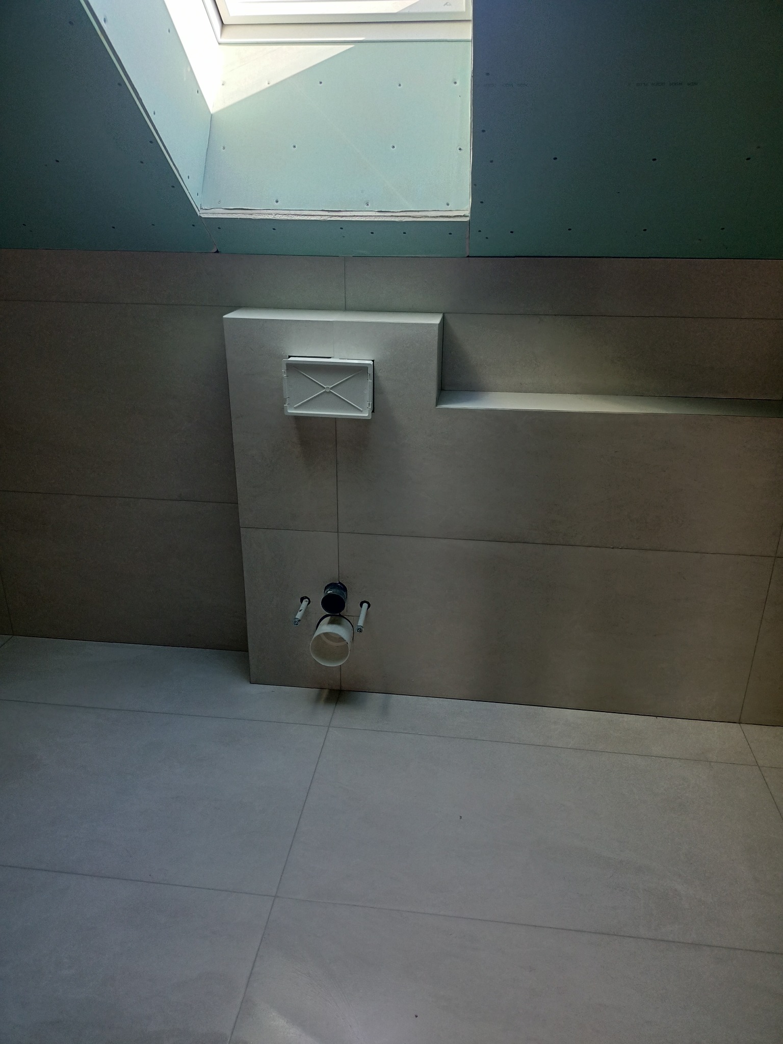 Szare płytki ceramiczne na ścianie i podłodze w łazience, widoczna instalacja podtynkowego WC i okno dachowe w suficie z płyt karton-gips.