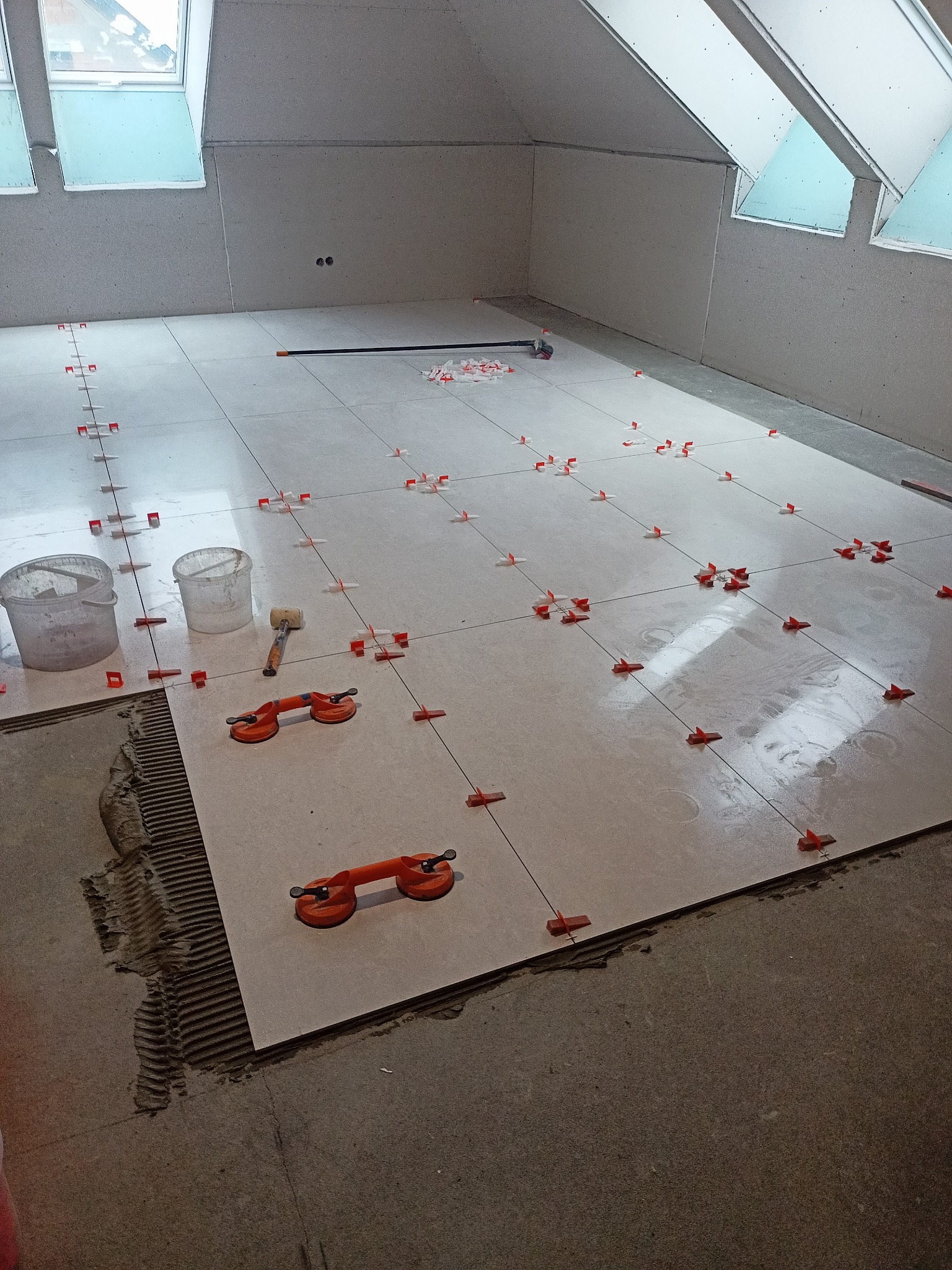 Układanie jasnych płytek podłogowych w pomieszczeniu na poddaszu z użyciem systemu poziomowania płytek; widoczne przyssawki do przenoszenia płytek, gumowy młotek i wiaderka.