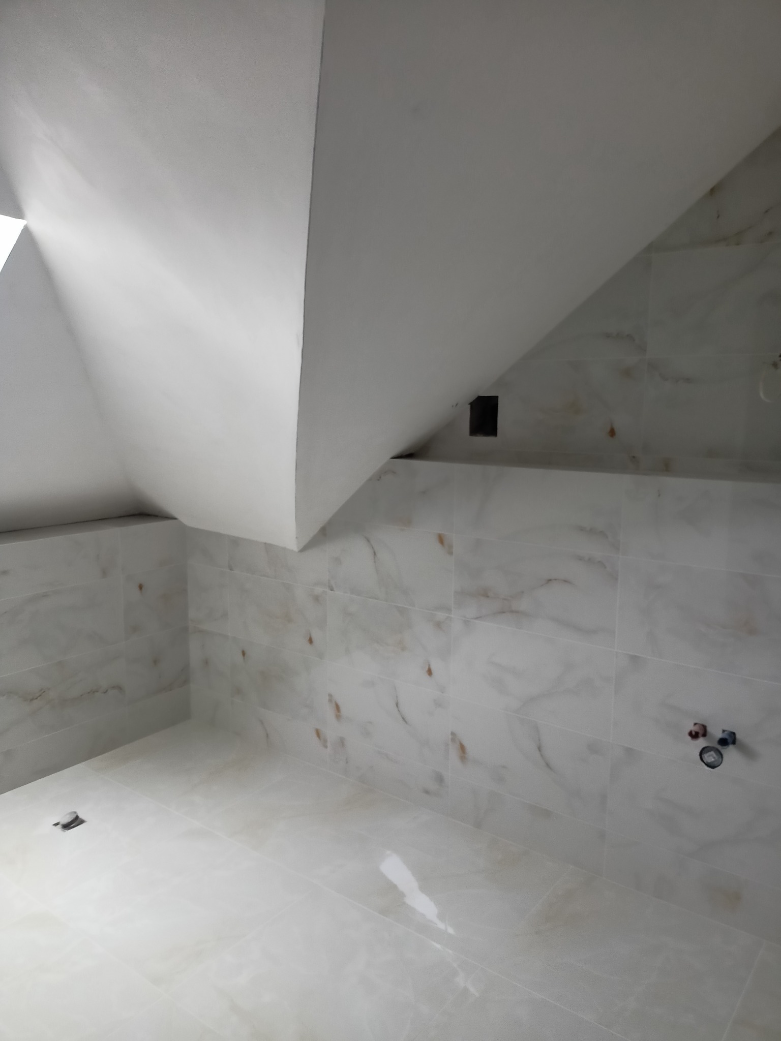 Wykończona łazienka na poddaszu ze skośnym sufitem, wyłożona białymi płytkami imitującymi marmur, widoczne przyłącza wodne i wentylacja.