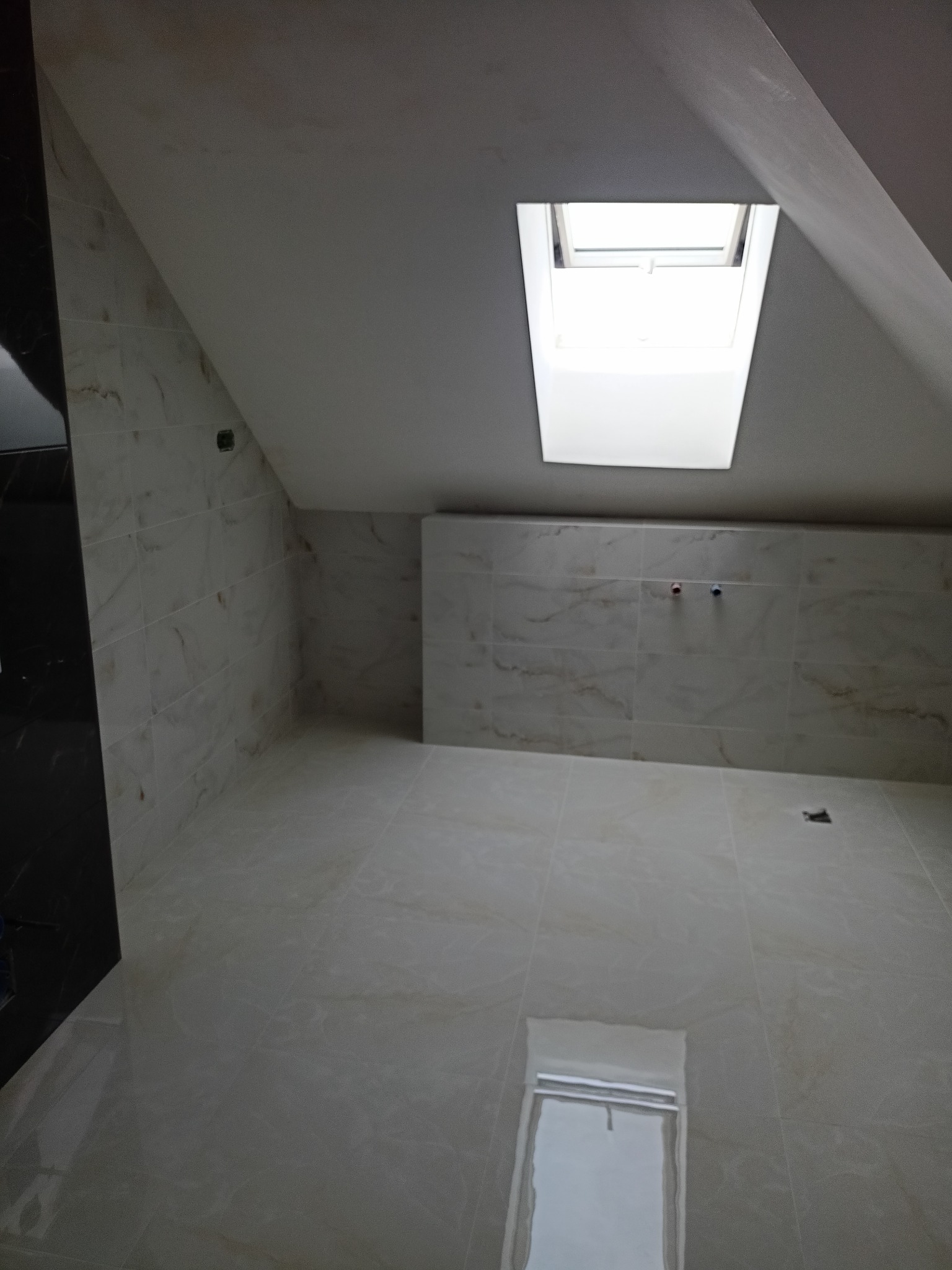 Wykończona łazienka na poddaszu z beżowymi płytkami imitującymi marmur, widoczne przyłącza wodne na ścianie i okno dachowe.