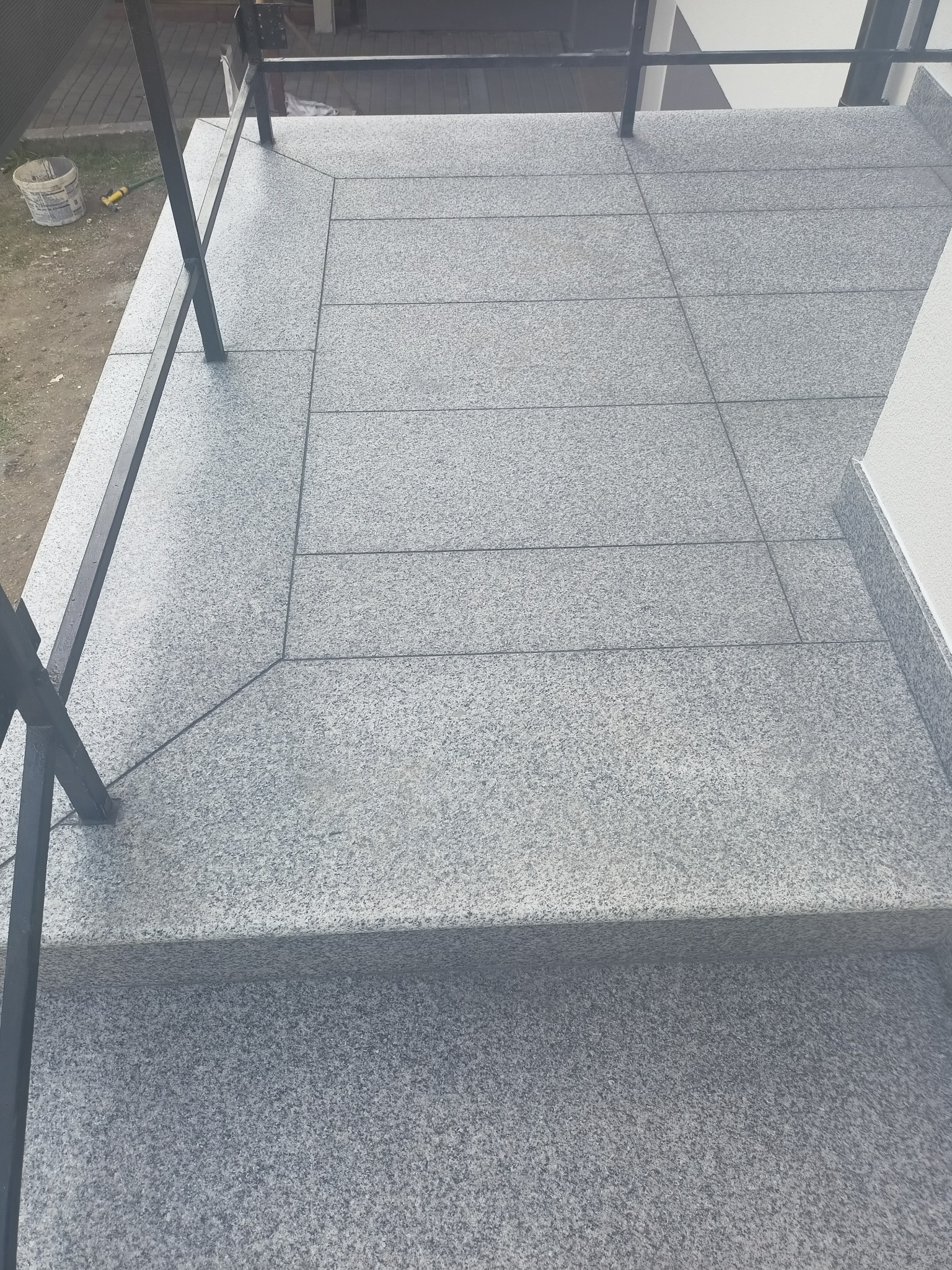 Wykończony taras w Rzeszowie z szarych, kwadratowych płytek, z czarną metalową barierką i widocznymi schodami wejściowymi.