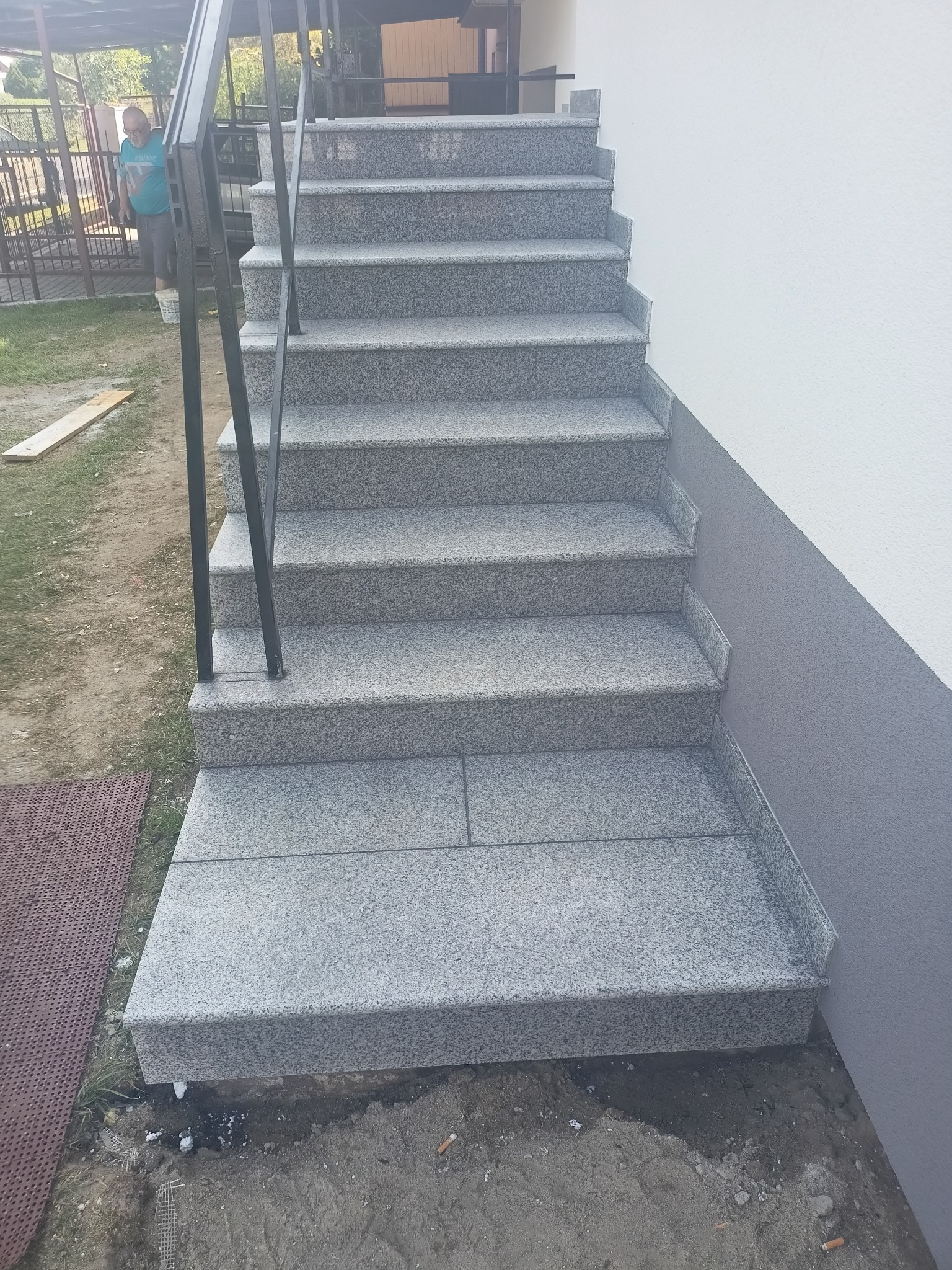 Zewnętrzne schody wyłożone jasnoszarym, nakrapianym granitem z czarną metalową barierką, prowadzące do budynku z biało-szarą elewacją.