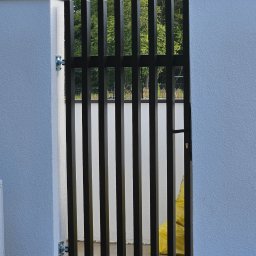 LUK-BUD Łukasz Wieteska - Nowoczesna brama wejściowa z pionowych, czarnych profili, zamontowana w białym murze w Dobrej. Minimalistyczny design, widok na zieleń w tle.
