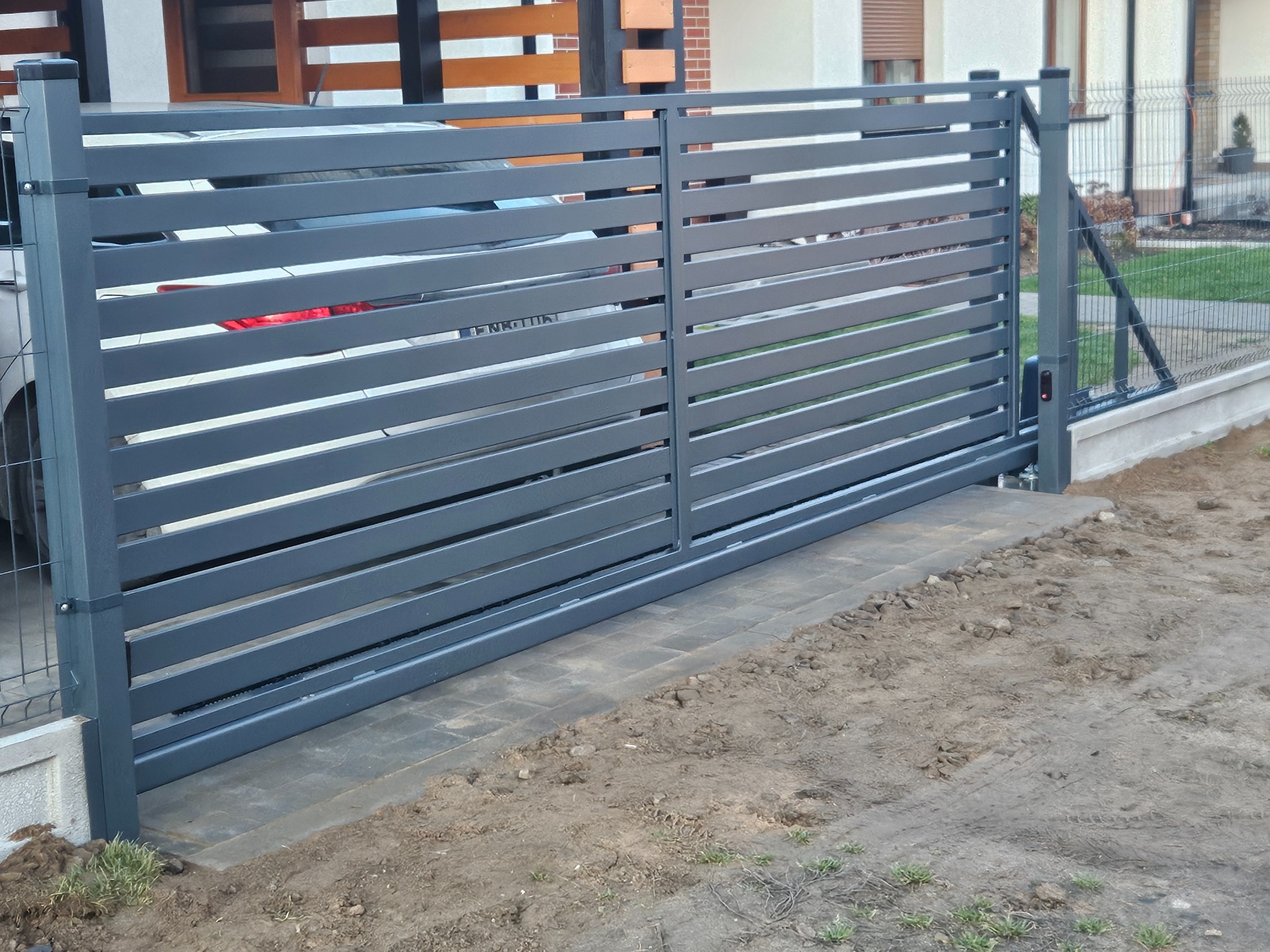 Nowoczesna, przesuwna brama wjazdowa z poziomych, szarych profili. Widoczny fragment samochodu za bramą i ogrodzenie panelowe po prawej stronie. Brama na kostce brukowej.