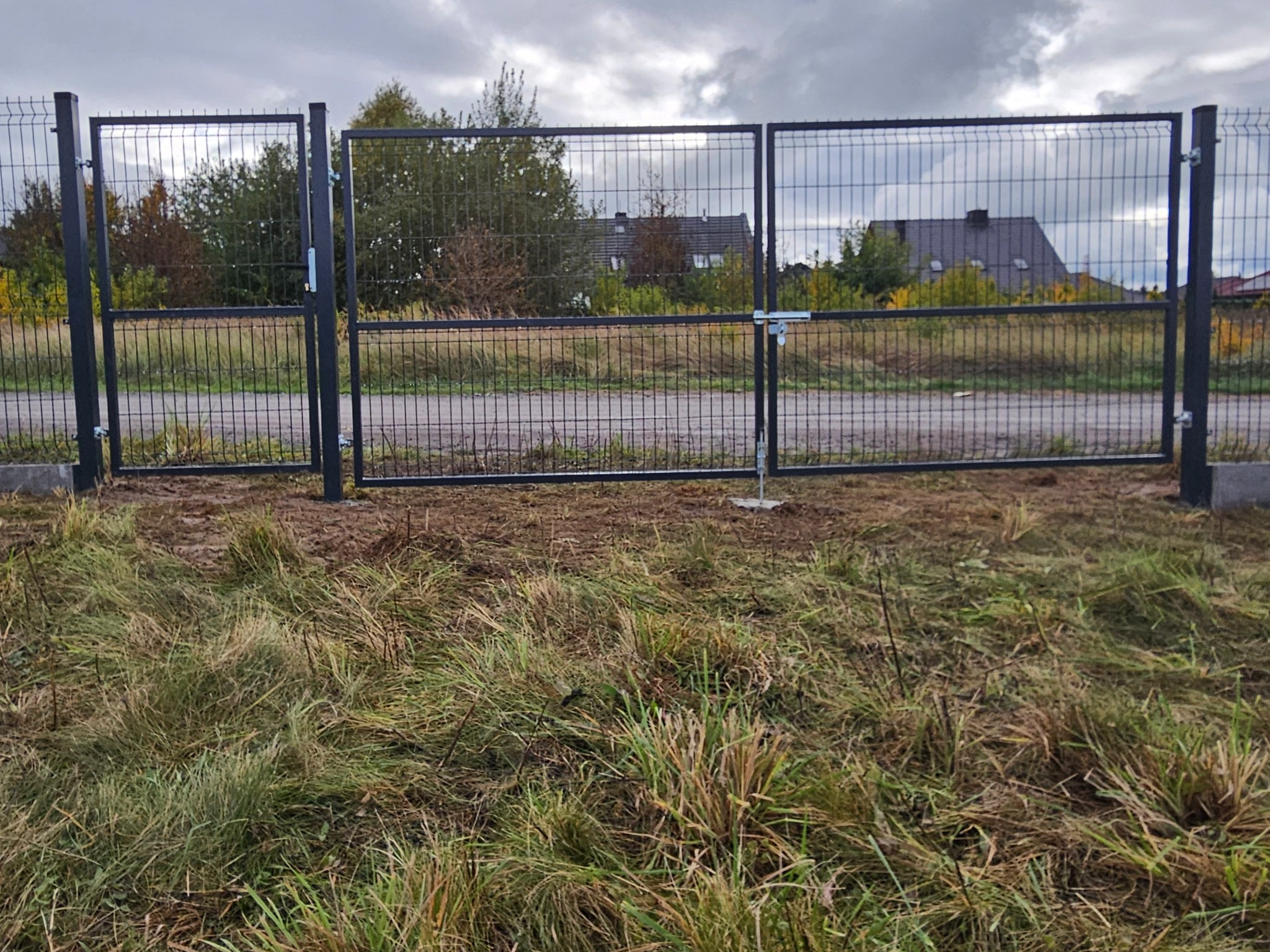 Czarna, metalowa brama panelowa z widocznym zamkiem na tle zieleni i zabudowań. Brama z siatki zgrzewanej, słupki zabetonowane w ziemi. Pochmurne niebo.