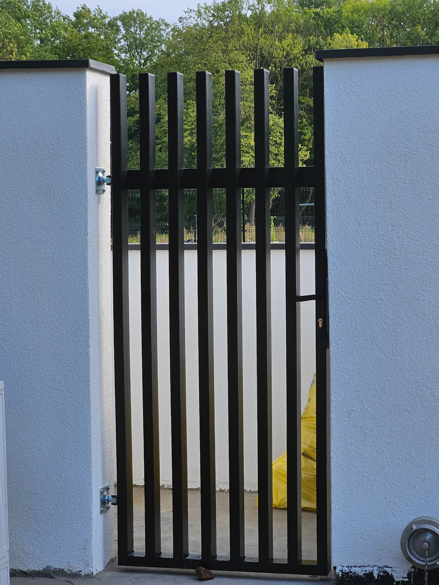 Nowoczesna brama wejściowa z pionowych, czarnych profili, zamontowana w białym murze w Dobrej. Minimalistyczny design, widok na zieleń w tle.