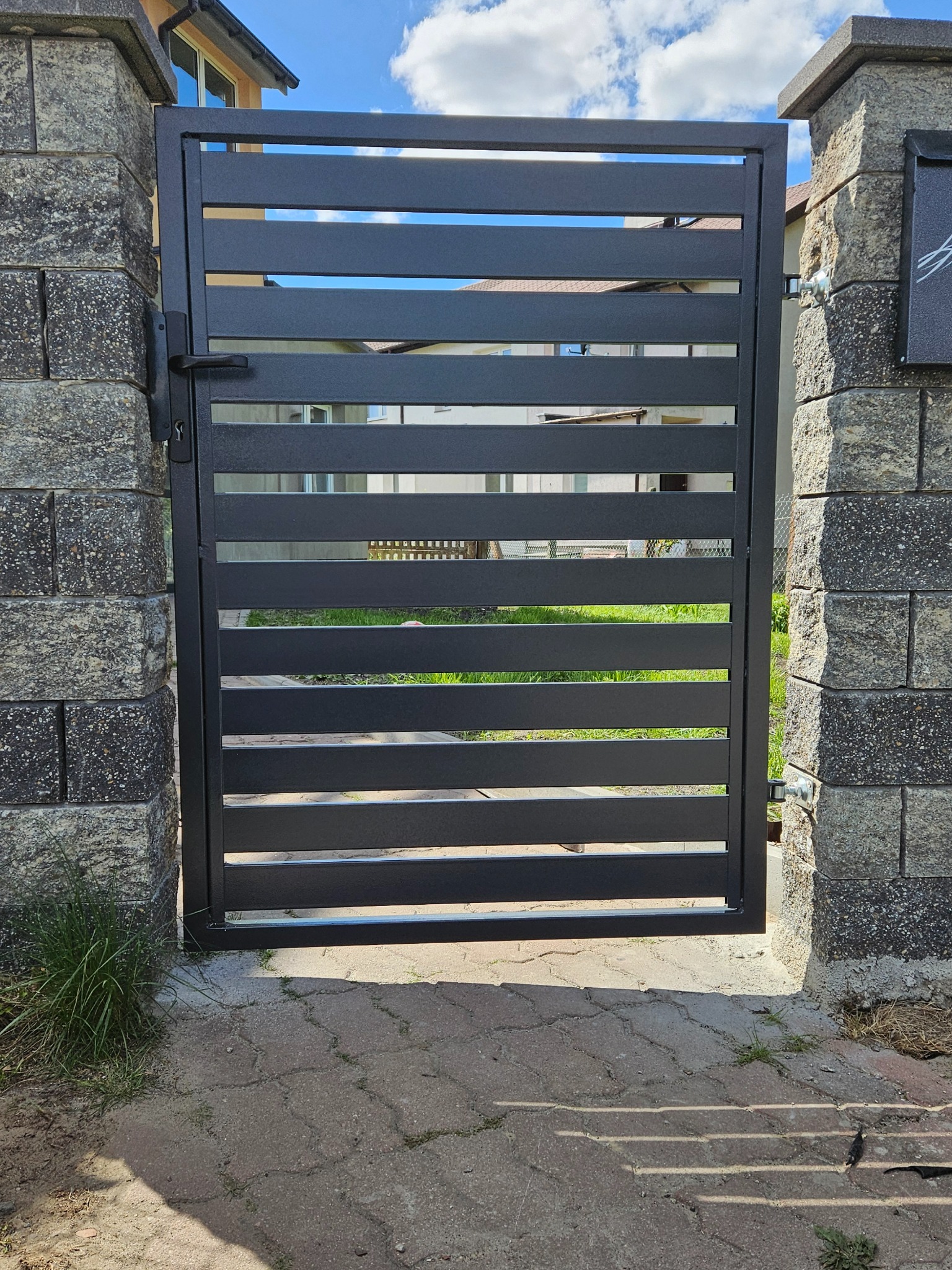 Nowoczesna, czarna brama wjazdowa z poziomymi panelami, zamontowana między kamiennymi słupkami. Widok na posesję przez prześwity między panelami.