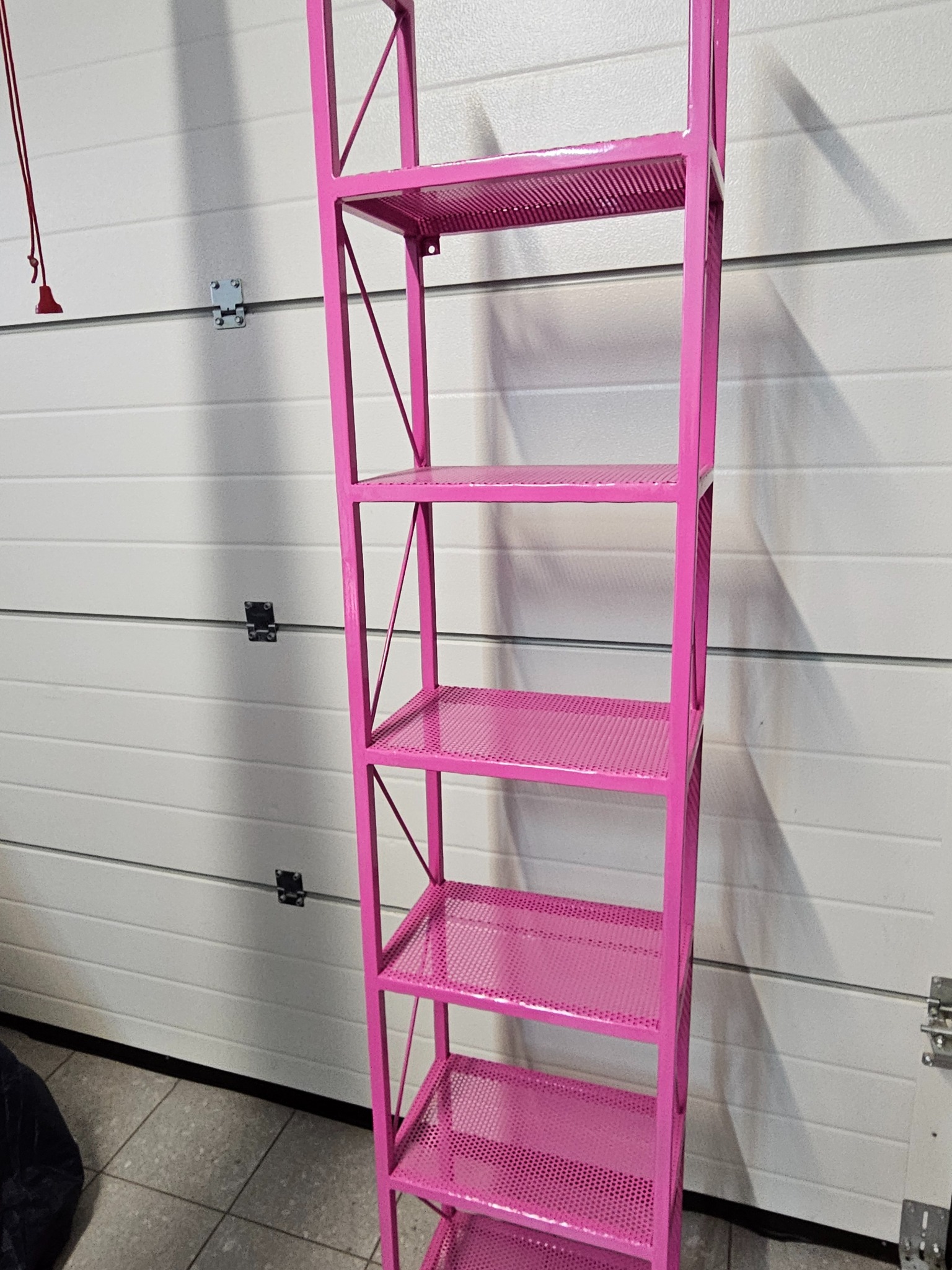 Różowa, metalowa konstrukcja regału z ażurowymi półkami na tle białej bramy garażowej. Nietypowy kolor i minimalistyczny design.