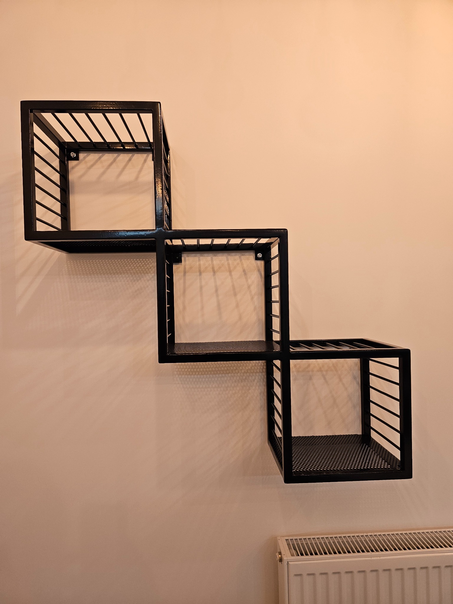 Czarna, metalowa półka ścienna w kształcie trzech połączonych kwadratów, zamocowana na jasnej ścianie. Widoczny fragment grzejnika.