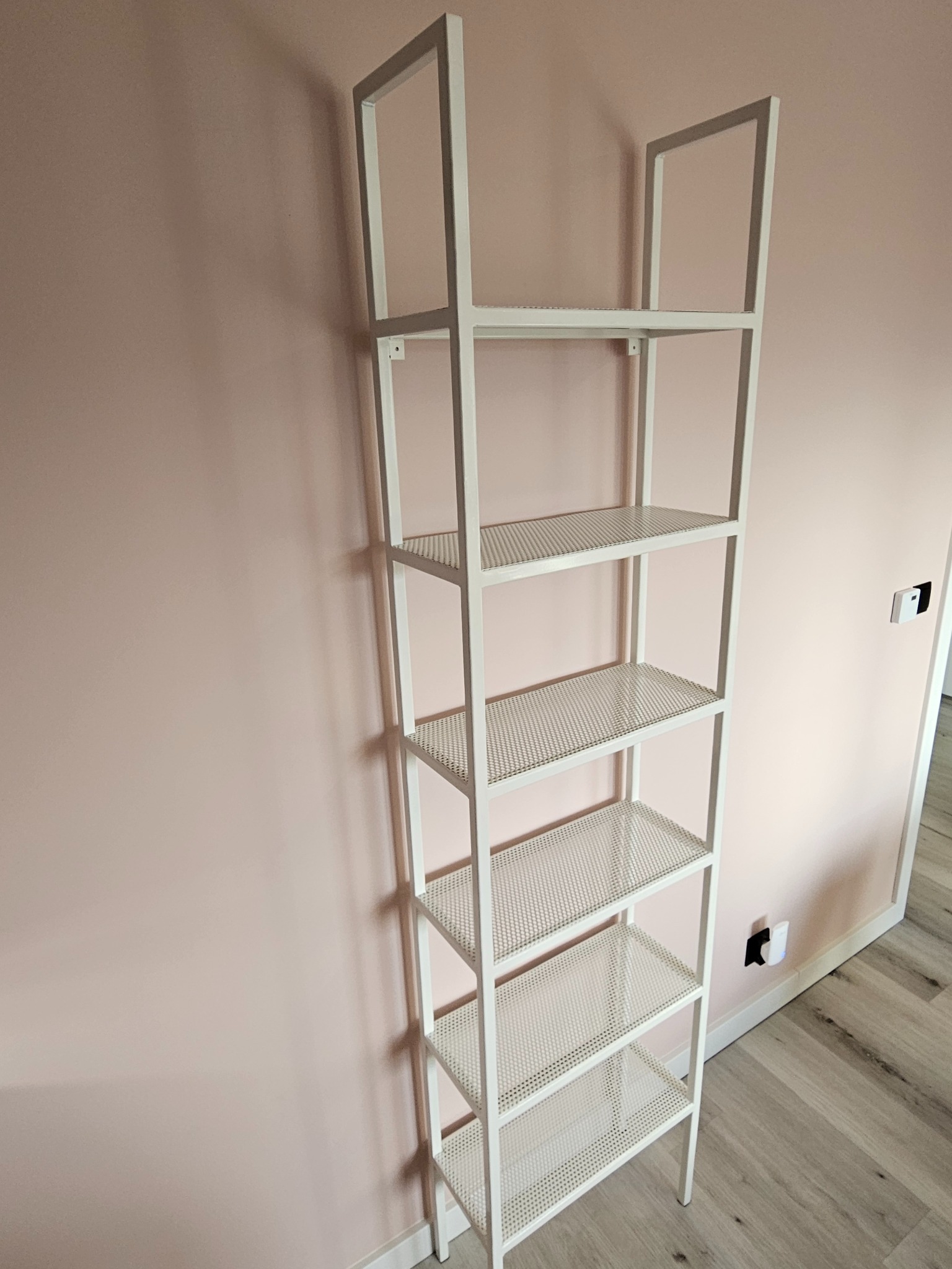 Biały, metalowy regał drabinkowy z ażurowymi półkami na tle pudrowo-różowej ściany i jasnej podłogi. Minimalistyczny design, pionowy kadr.