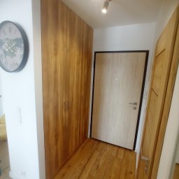 Michał Wojnar Jawor Meble - Wnętrze przedpokoju z drewnianą podłogą, szafą wnękową z drewnianymi frontami i drzwiami wejściowymi. Na ścianie widoczny zegar. Oświetlenie punktowe.