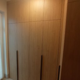 Michał Wojnar Jawor Meble - Drewniana szafa wnękowa z długimi, pionowymi uchwytami, oświetlona ciepłym światłem, z widocznym fragmentem okna z żaluzjami po lewej stronie.