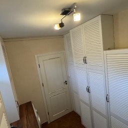 Michał Wojnar Jawor Meble - Białe szafy z lamelowymi drzwiami, ustawione wzdłuż ściany, oświetlone przez reflektory sufitowe. Widoczna również biała, klasyczna stolarka drzwiowa.