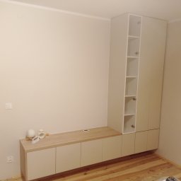 Michał Wojnar Jawor Meble - Jasna zabudowa meblowa z szafkami i półkami, z drewnianym blatem, na tle beżowej ściany i drewnianej podłogi. Minimalistyczny design, widoczne detale wykończenia.