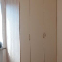 Michał Wojnar Jawor Meble - Czterodrzwiowa szafa wnękowa z jasnego drewna i beżowymi frontami, z delikatnymi, srebrnymi uchwytami, na drewnianej podłodze. Widok na narożnik szafy.