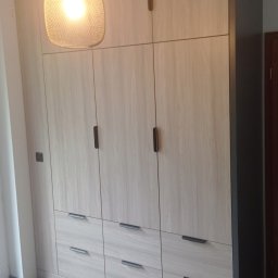 Michał Wojnar Jawor Meble - Jasna, drewnopodobna szafa wnękowa z ciemnymi uchwytami, oświetlona wiszącą lampą. Szare wykończenie krawędzi. Widoczne panele podłogowe.