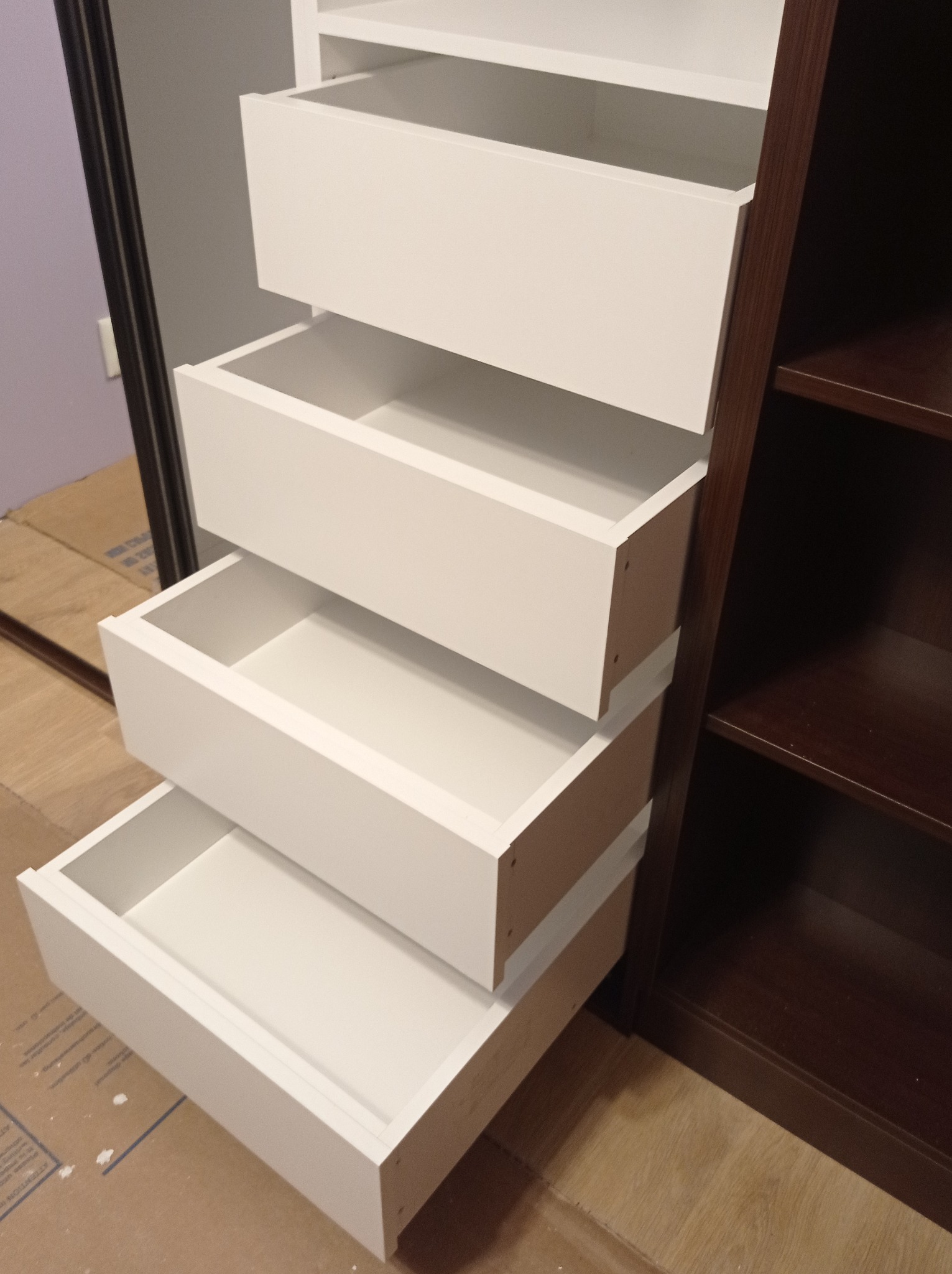 Otwarta szafa wnękowa z czterema wysuniętymi, białymi szufladami, widoczne brązowe półki i karton na podłodze.