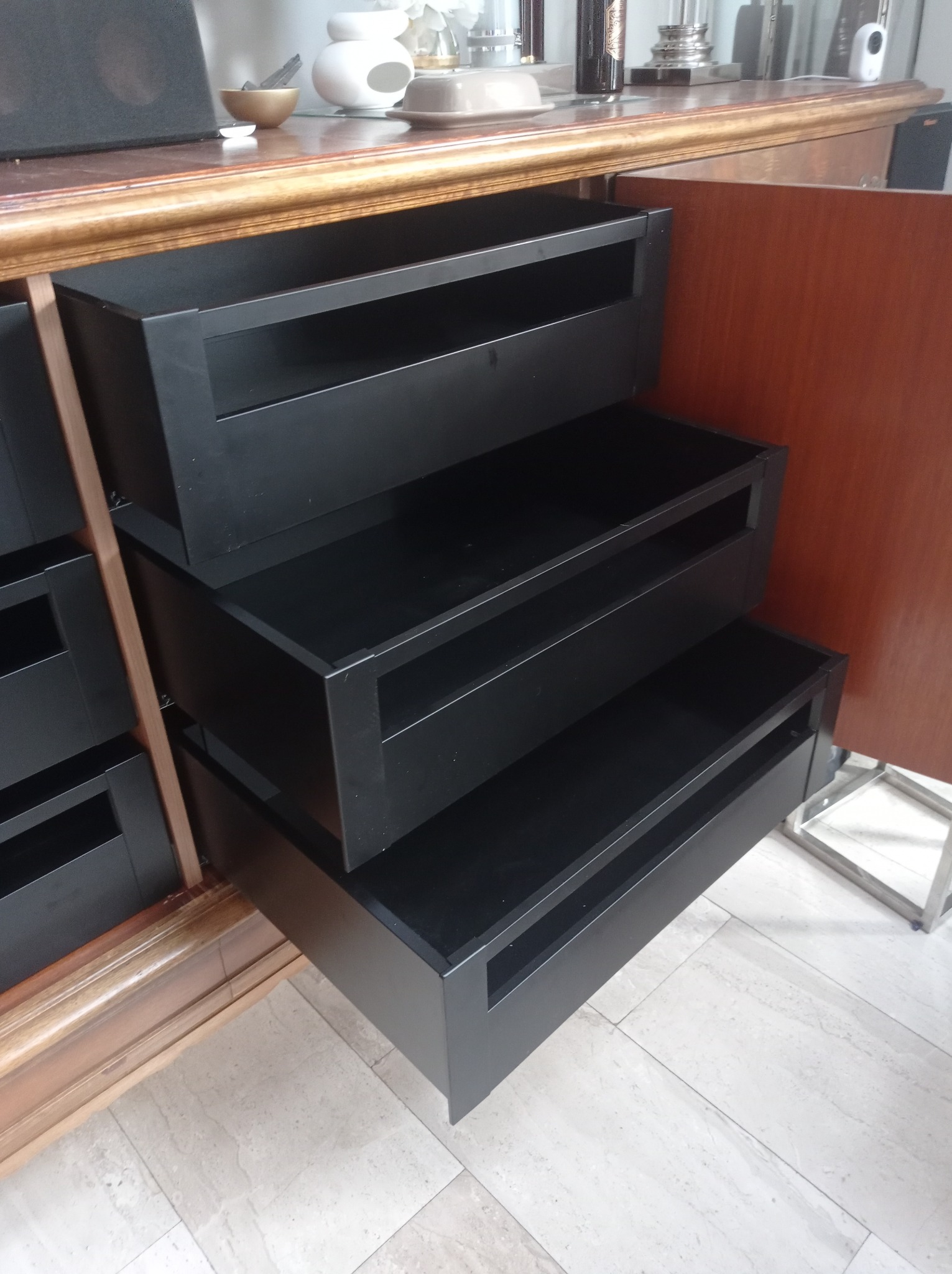 Wysuwane czarne szuflady z metalowymi ramkami w szafie o drewnianej obudowie z dekoracyjnymi listwami.