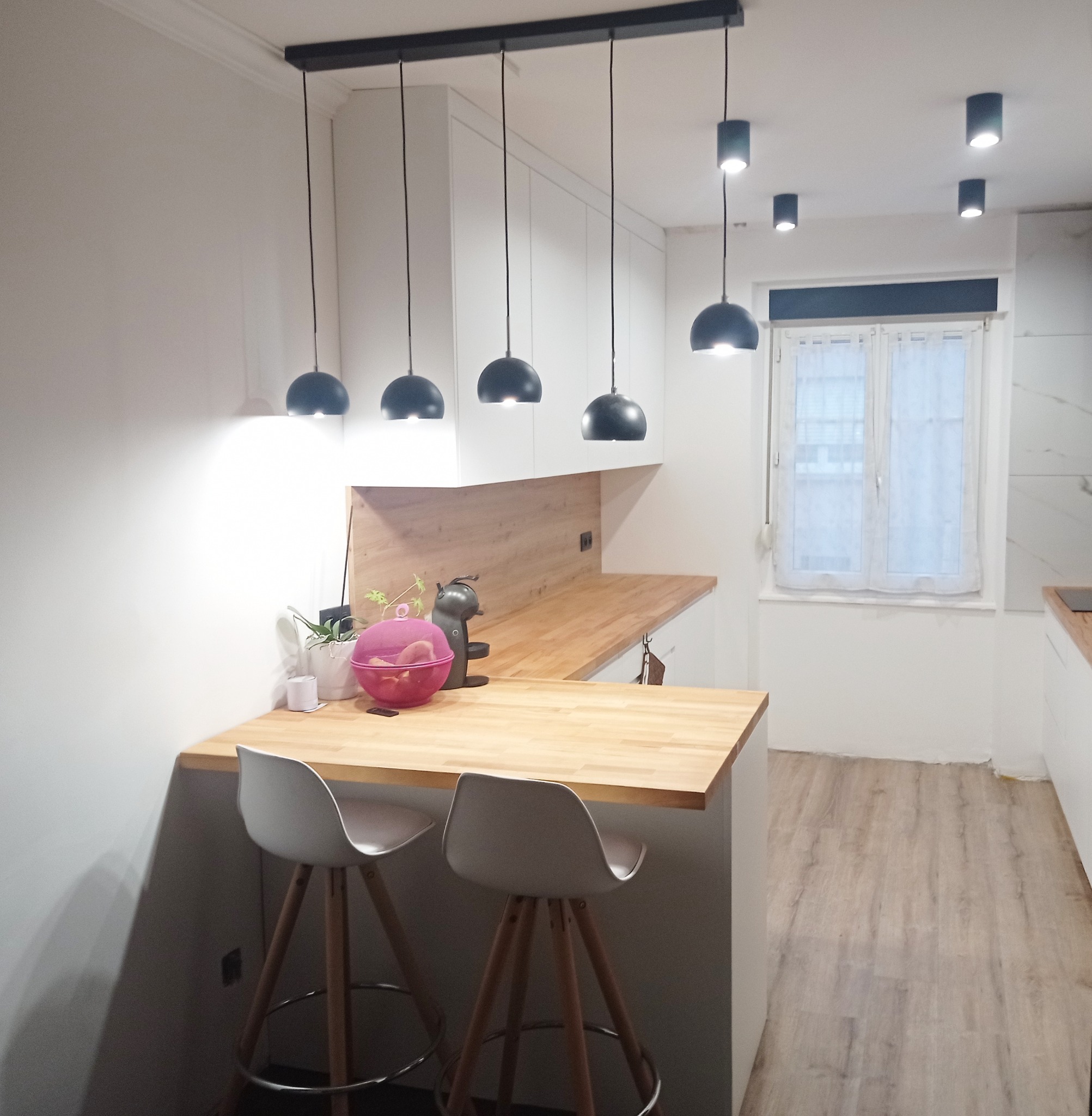 Nowoczesna kuchnia z białymi szafkami, drewnianym blatem, wyspą z dwoma białymi hokerami i czarnymi lampami wiszącymi oraz punktowym oświetleniem sufitowym.