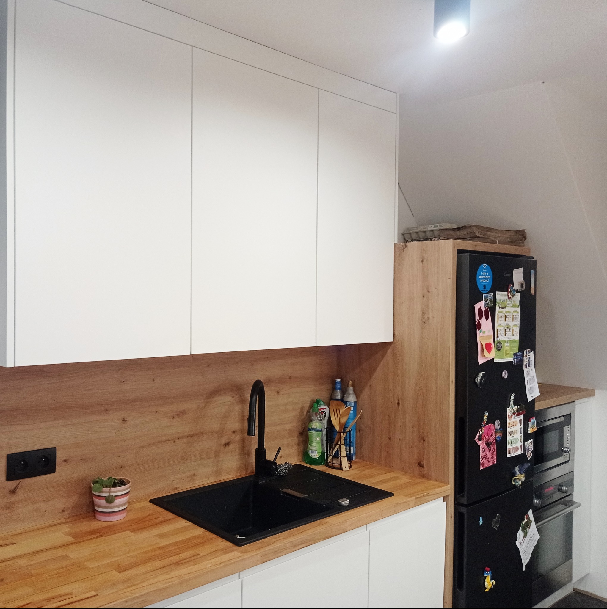 Nowoczesna kuchnia z białymi szafkami, drewnianym blatem i czarnym zlewem, uzupełniona o czarną lodówkę z magnesami i drewniane wykończenie ściany.