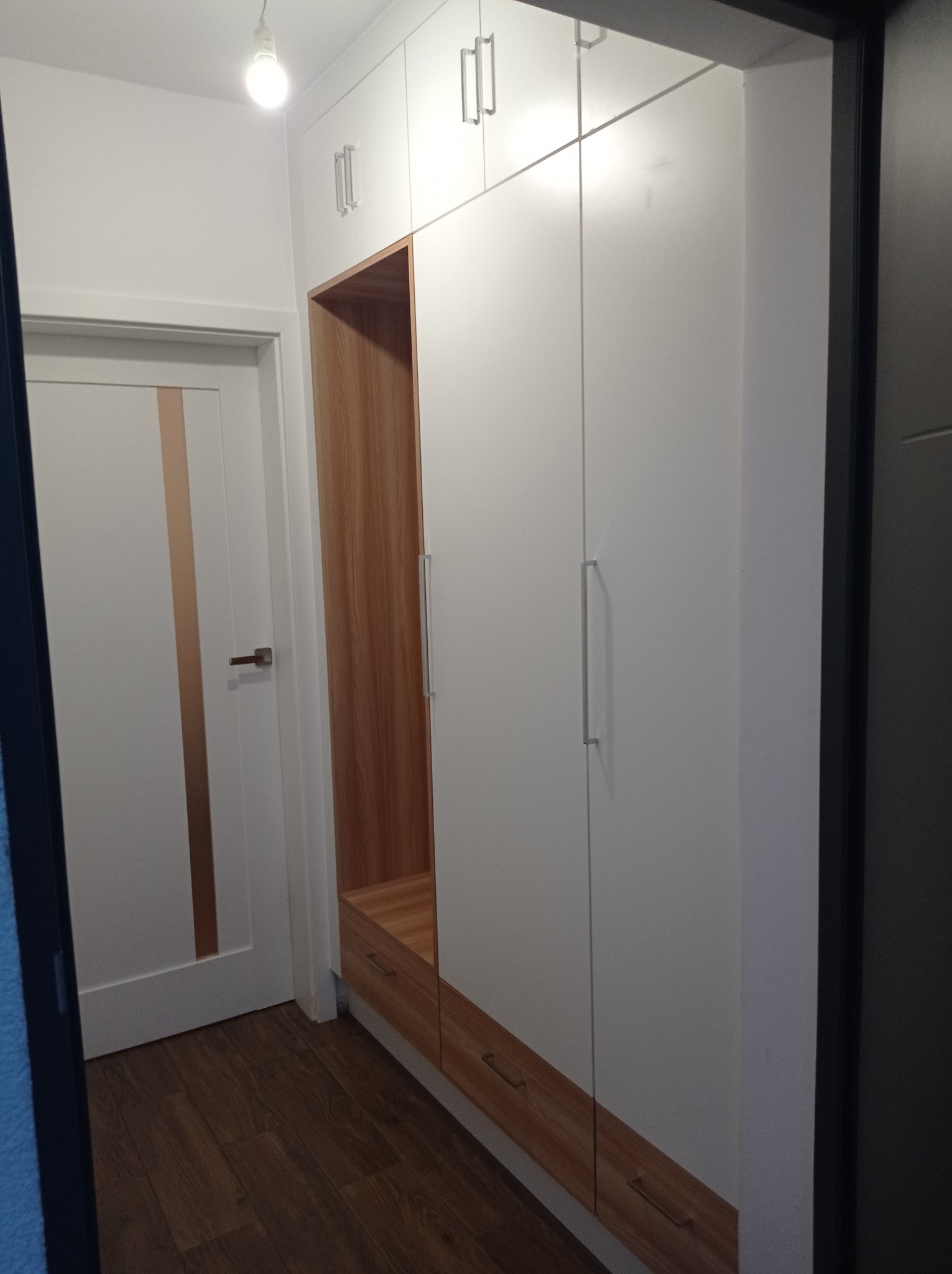Biała szafa wnękowa z drewnianymi akcentami i otwartą przestrzenią na buty, umieszczona w przedpokoju obok białych drzwi z drewnianym paskiem.