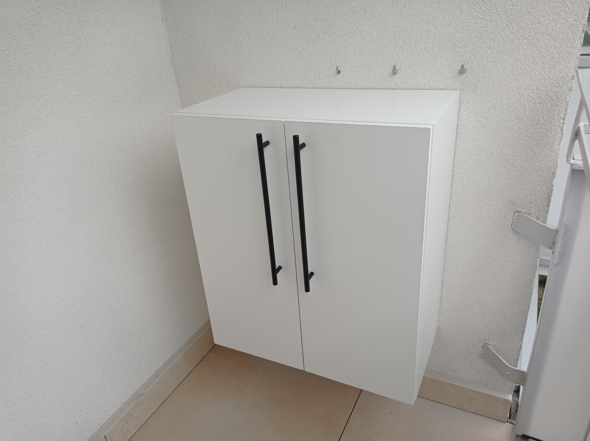Biała szafka na wymiar z czarnymi, podłużnymi uchwytami zamontowana na ścianie o szorstkiej fakturze, z widocznymi haczykami powyżej.