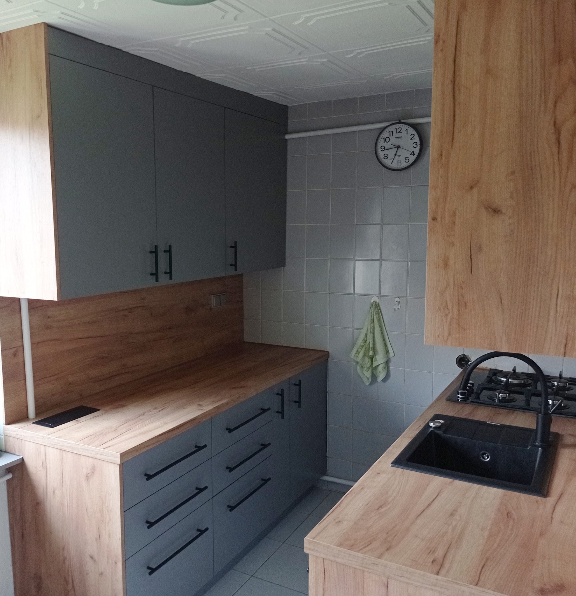 Szare szafki kuchenne z drewnianymi blatami i czarnymi uchwytami, widoczny zlew, płyta gazowa i zegar na ścianie z płytek.