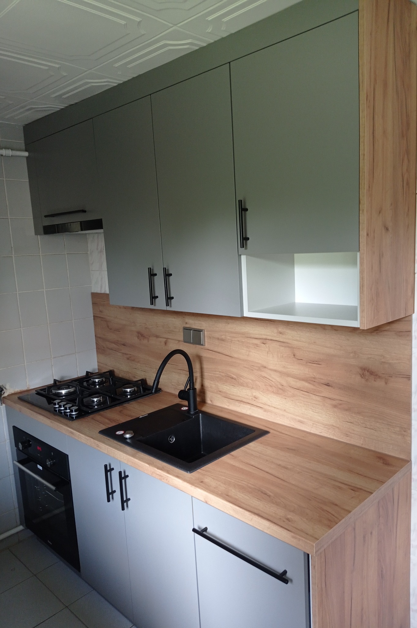 Szare szafki kuchenne z drewnianym blatem, czarny zlew i bateria, płyta gazowa, piekarnik, biała podsufitka z wzorem.
