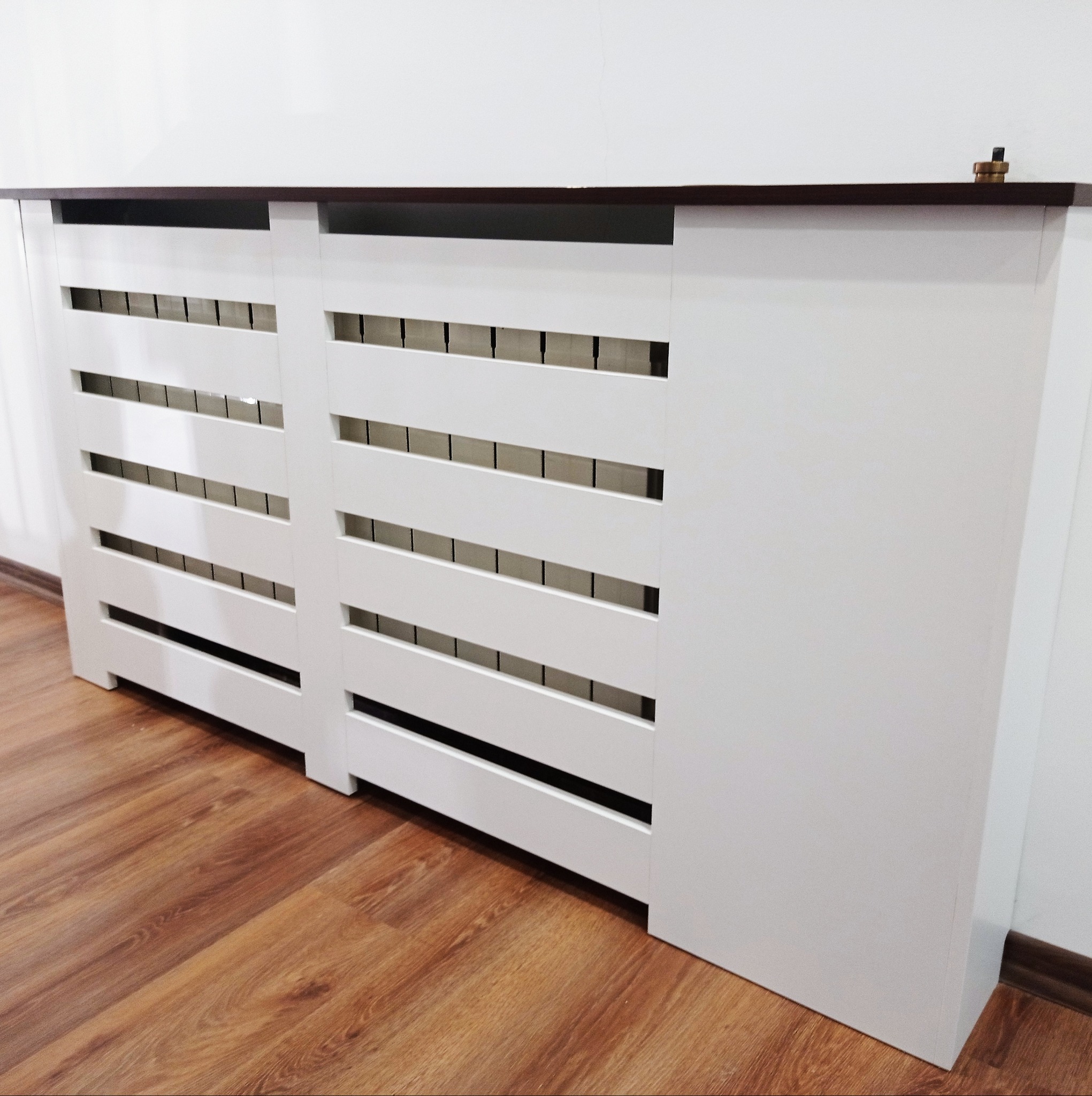 Biała obudowa grzejnika z poziomymi szczelinami wentylacyjnymi i ciemnobrązowym blatem, zamontowana na drewnianej podłodze przy białej ścianie, widoczny zawór grzejnika.