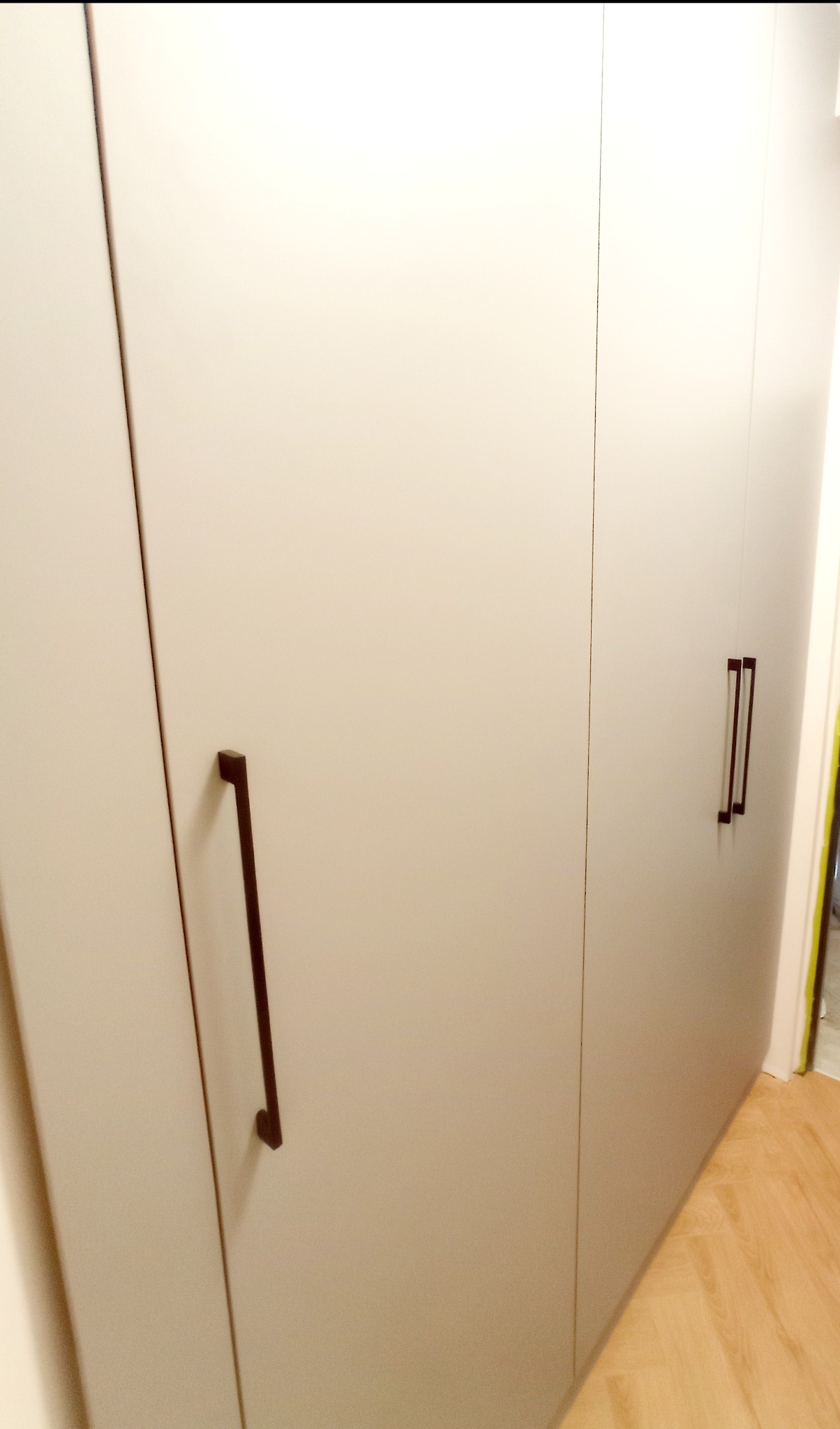 Jasnoszara szafa wnękowa z długimi, prostokątnymi uchwytami w kolorze ciemnego brązu, widoczna z boku na tle drewnianej podłogi.