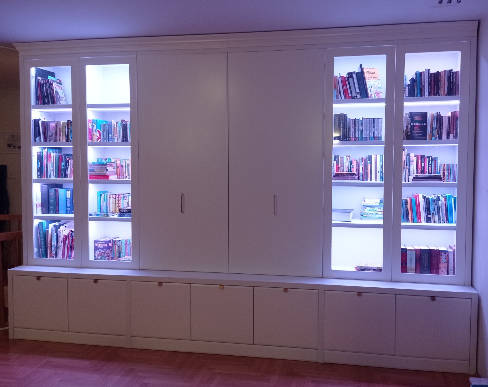 Biała zabudowa meblowa z przeszklonymi witrynami podświetlonymi LED, pełnymi książek, oraz szafkami dolnymi z minimalistycznymi uchwytami.