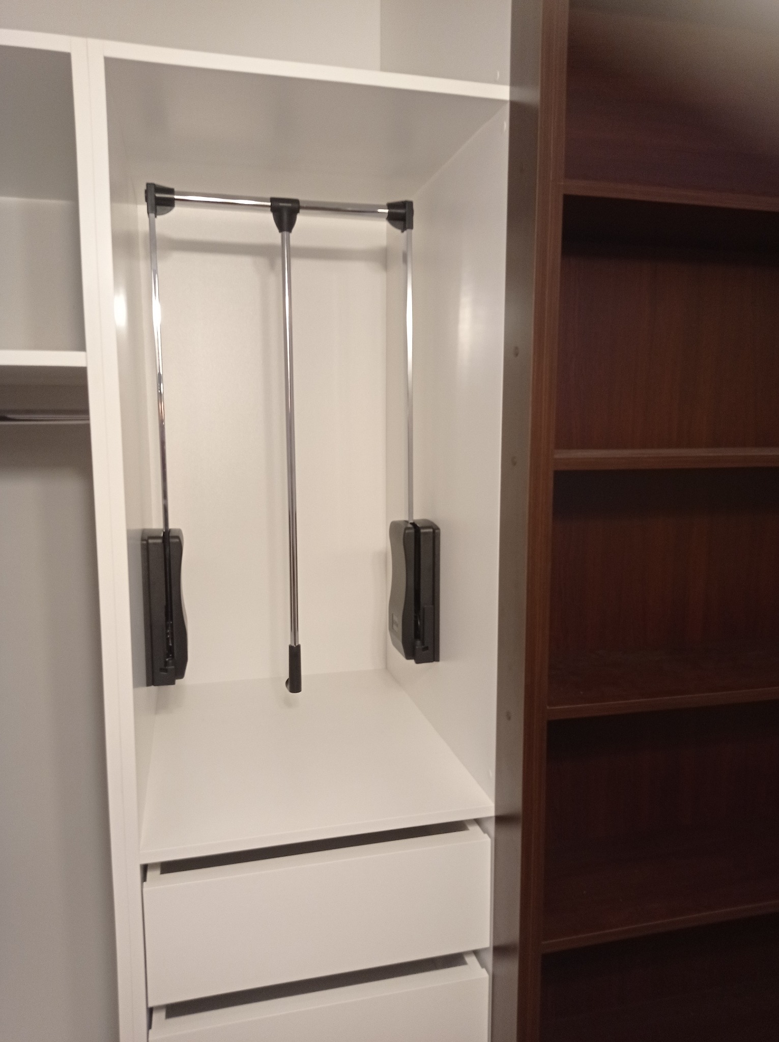 Wnętrze szafy z jasnymi frontami szuflad, wyposażone w system drążków opuszczanych do wieszania ubrań, widoczna również część brązowej szafy z półkami.