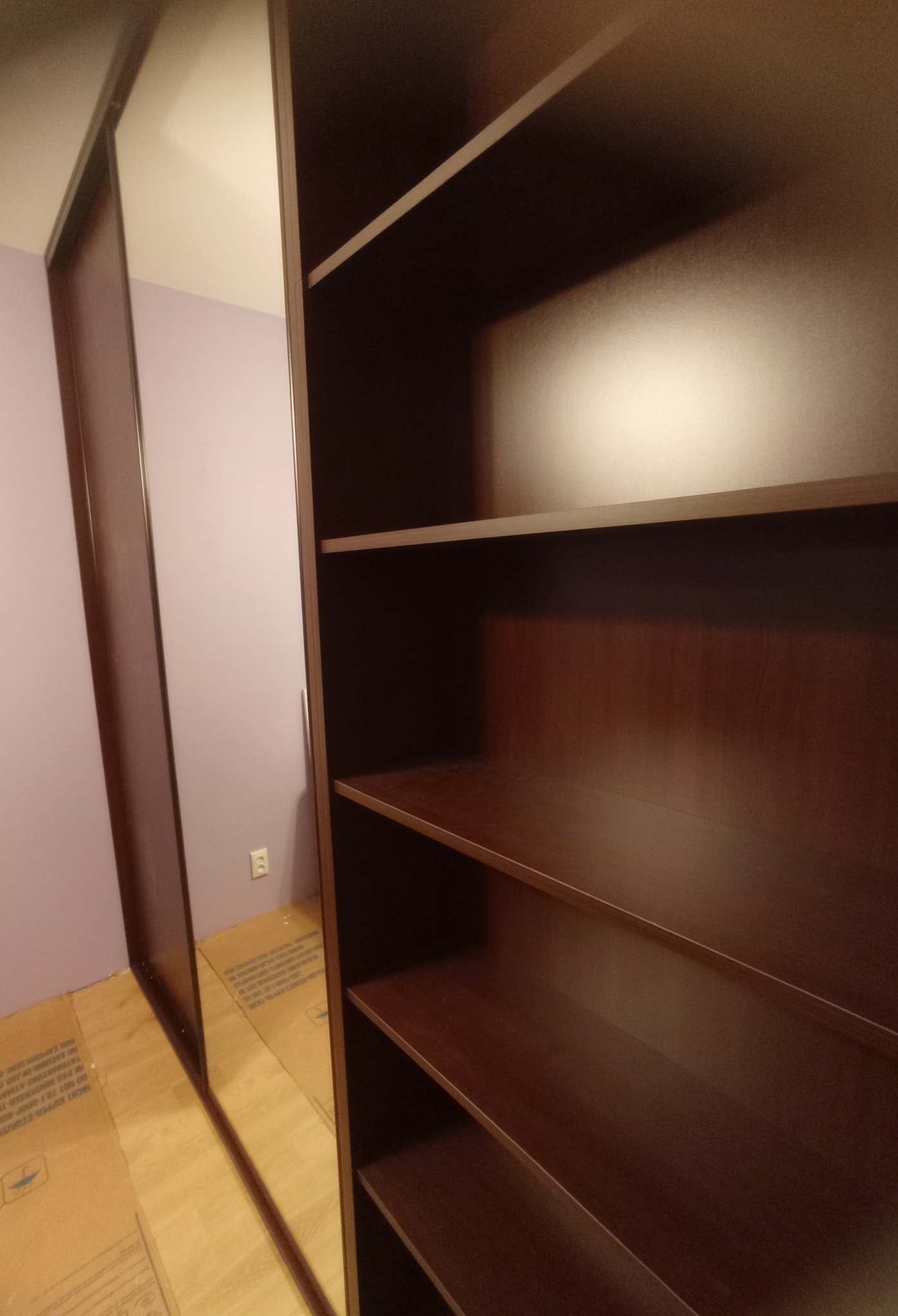 Ciemnobrązowa szafa wnękowa z przesuwnymi drzwiami lustrzanymi, częściowo pusta, na podłodze zabezpieczonej kartonem, widok z boku.