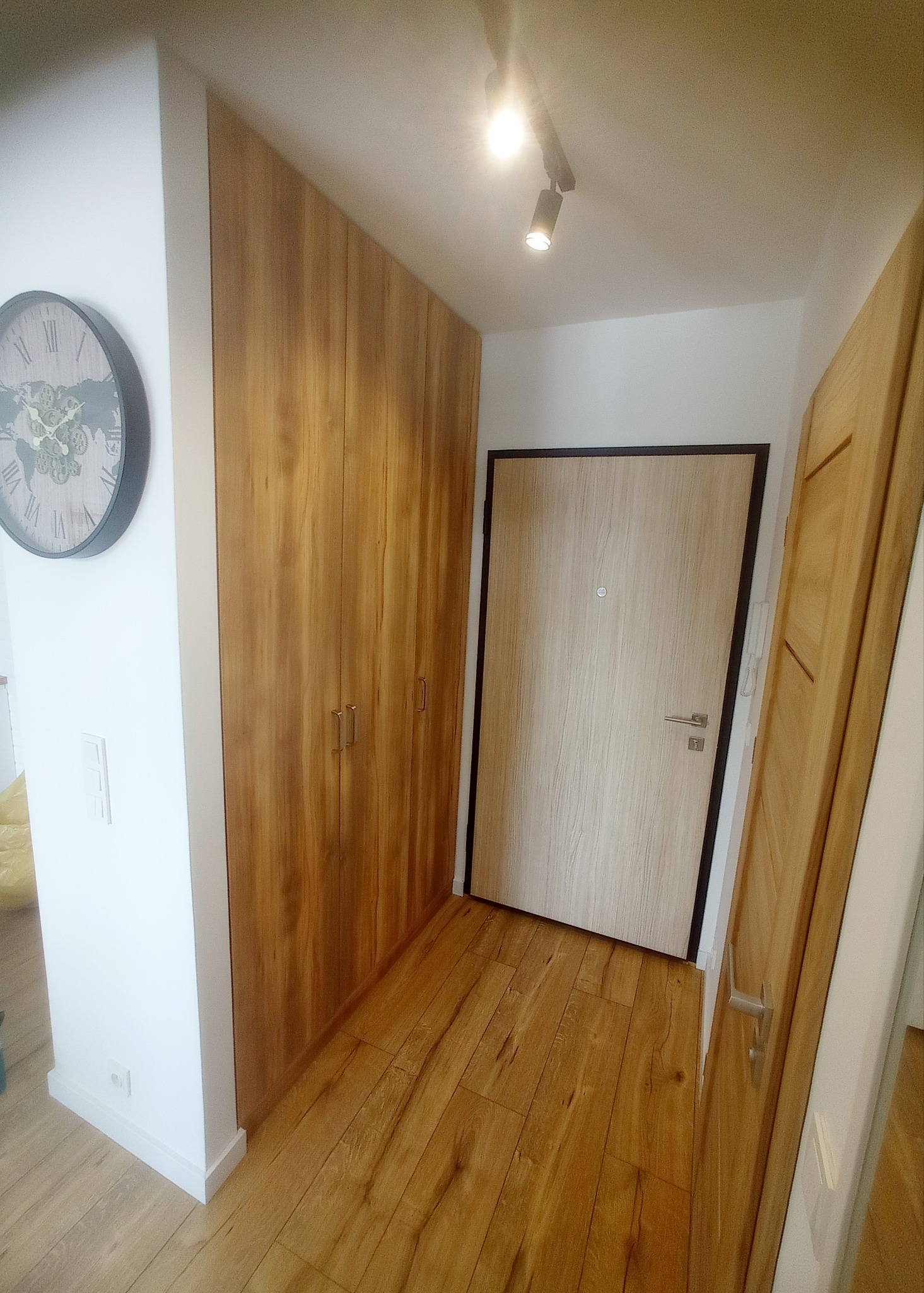 Wnętrze przedpokoju z drewnianą podłogą, szafą wnękową z drewnianymi frontami i drzwiami wejściowymi. Na ścianie widoczny zegar. Oświetlenie punktowe.