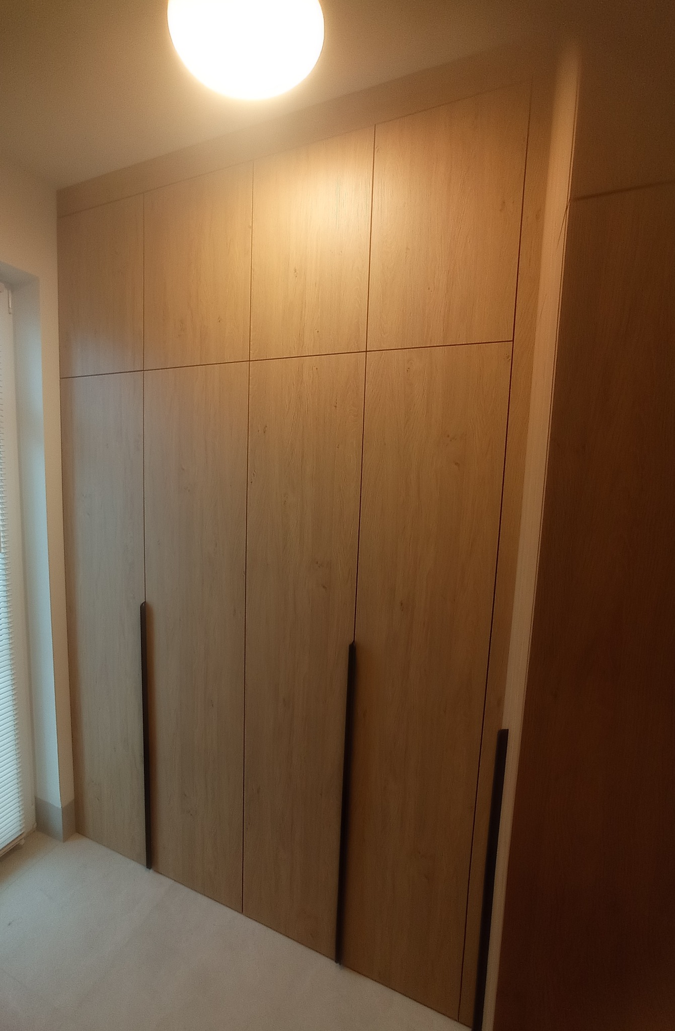 Drewniana szafa wnękowa z długimi, pionowymi uchwytami, oświetlona ciepłym światłem, z widocznym fragmentem okna z żaluzjami po lewej stronie.