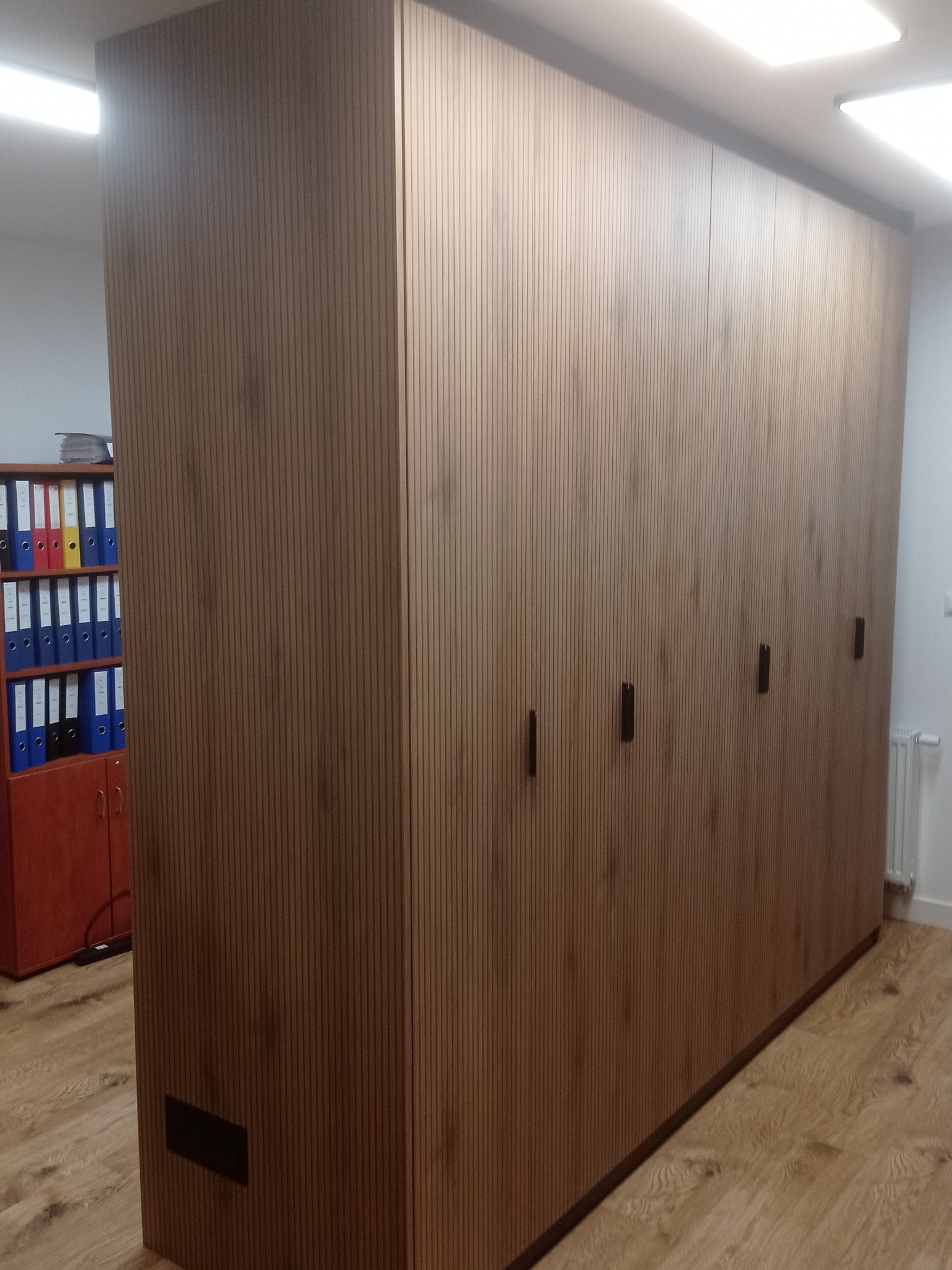 Duża, brązowa szafa wnękowa z pionowymi żłobieniami i minimalistycznymi uchwytami, stojąca na drewnianej podłodze. W tle regał z segregatorami.