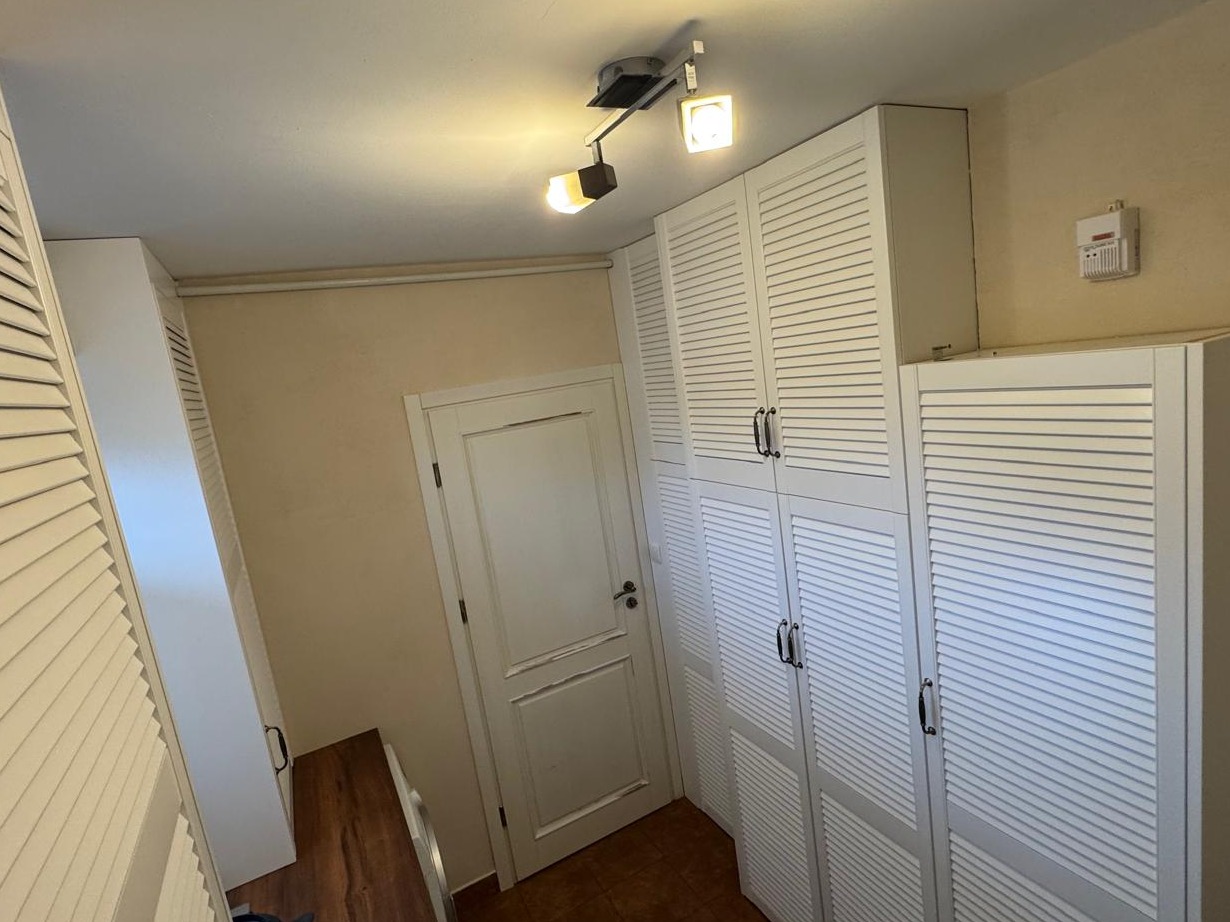 Białe szafy z lamelowymi drzwiami, ustawione wzdłuż ściany, oświetlone przez reflektory sufitowe. Widoczna również biała, klasyczna stolarka drzwiowa.