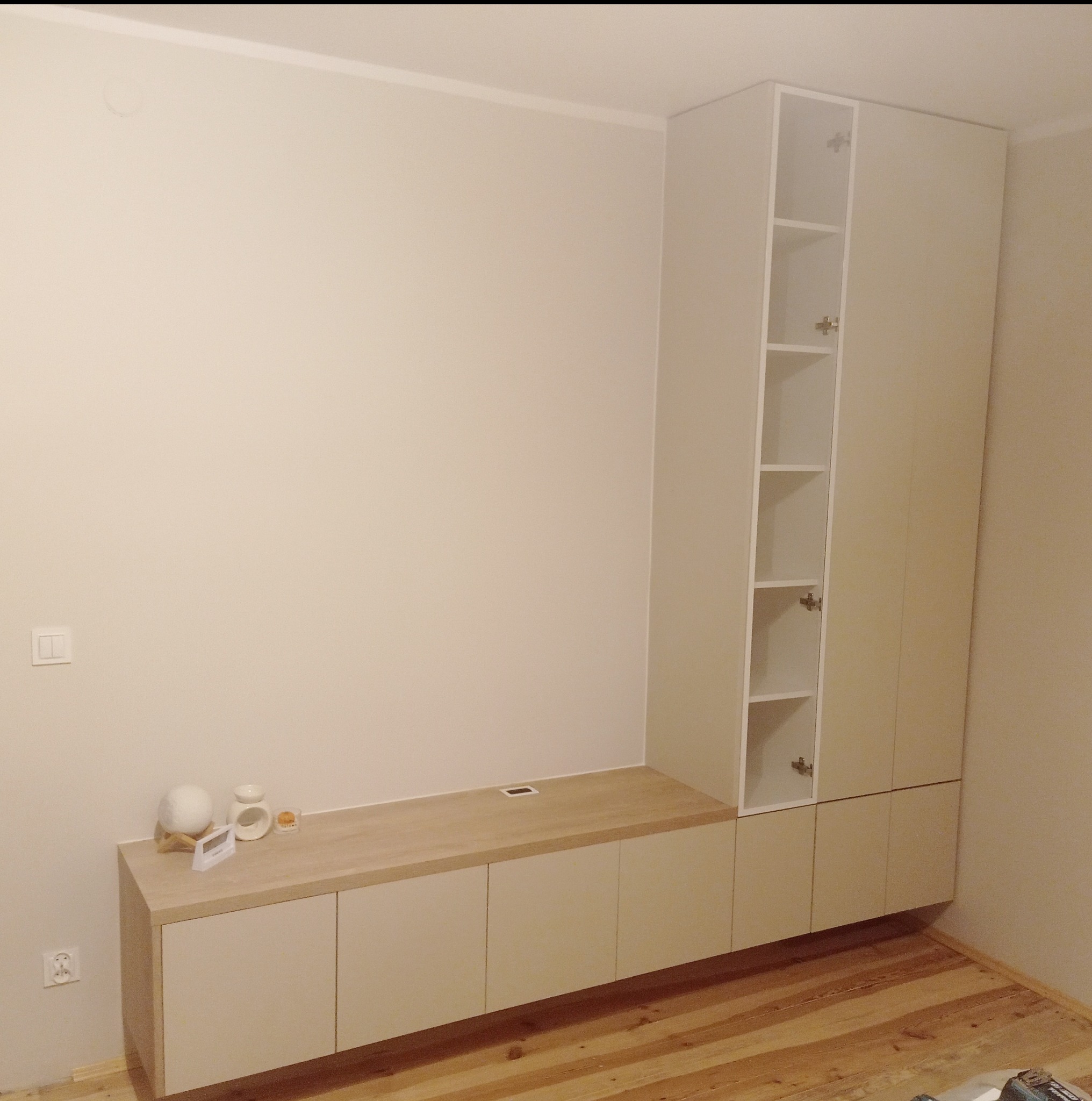 Jasna zabudowa meblowa z szafkami i półkami, z drewnianym blatem, na tle beżowej ściany i drewnianej podłogi. Minimalistyczny design, widoczne detale wykończenia.