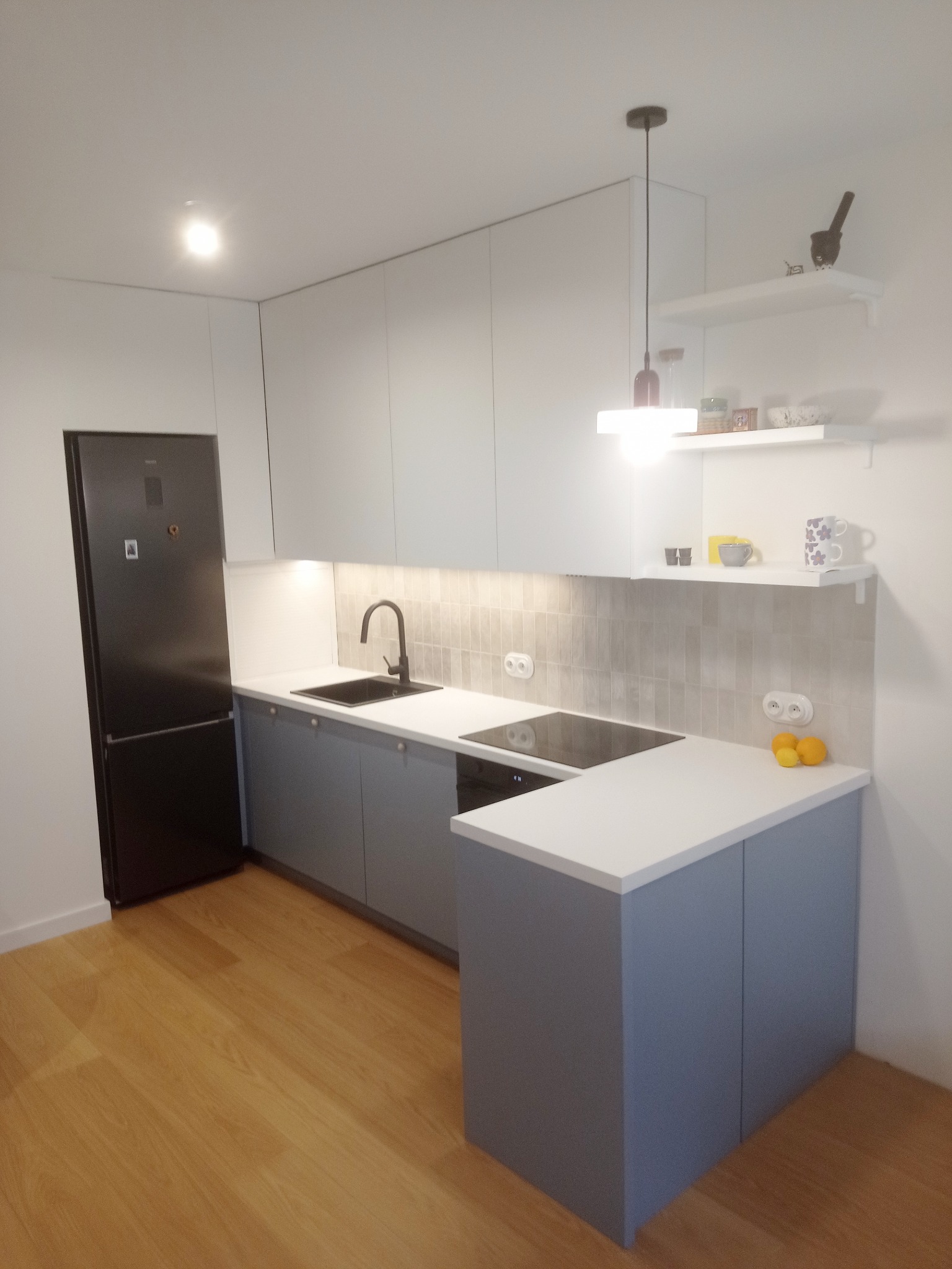 Nowoczesna kuchnia z szarymi szafkami dolnymi i białymi górnymi, czarnym zlewem i płytą indukcyjną. Na ścianie półki z dekoracjami. Drewniana podłoga.